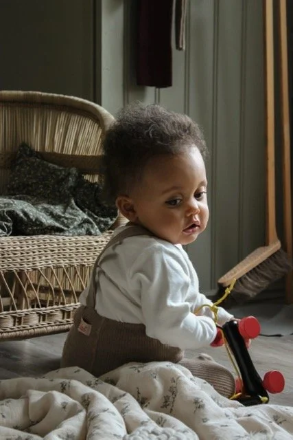 A young child sitting on the floor next to a stuffed blanket, holding a wooden toy with red wheels in a cozy indoor setting.