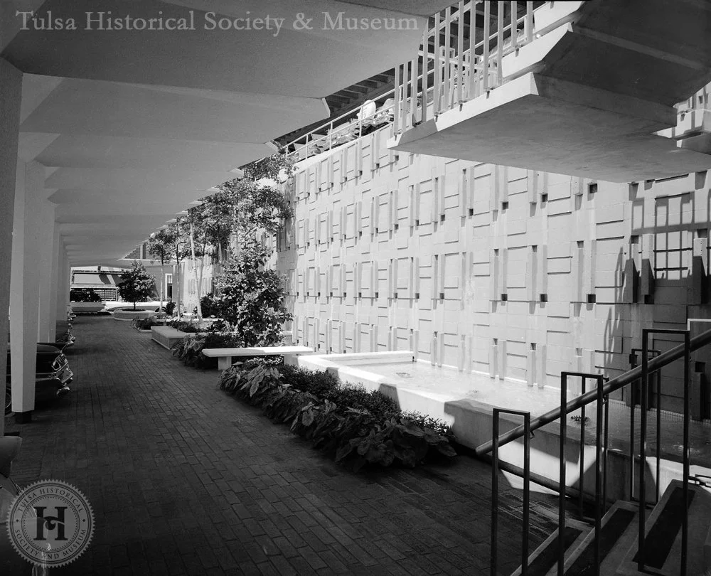 Modern exterior corridor with patterned wall, landscaped plants, benches, parked cars, and a stairway with railing outside Tulsa Historical Society and Museum.
