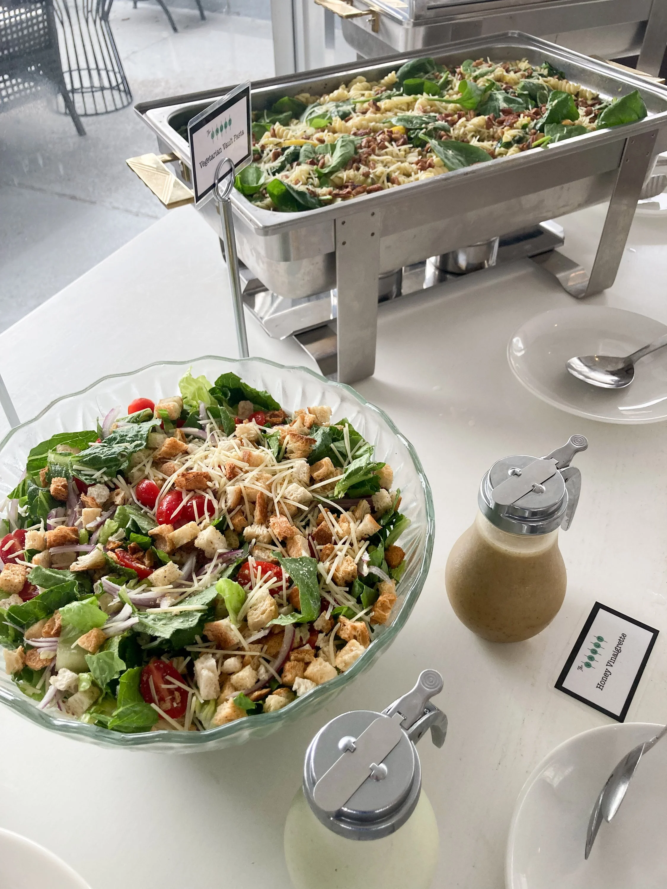 A large glass bowl of salad containing greens, cherry tomatoes, shredded cheese, croutons, and chopped vegetables, labeled 'Honey Vinaigrette' on a stand. In the background, a chafing dish with pasta salad and a bottle of salad dressing on a white ta