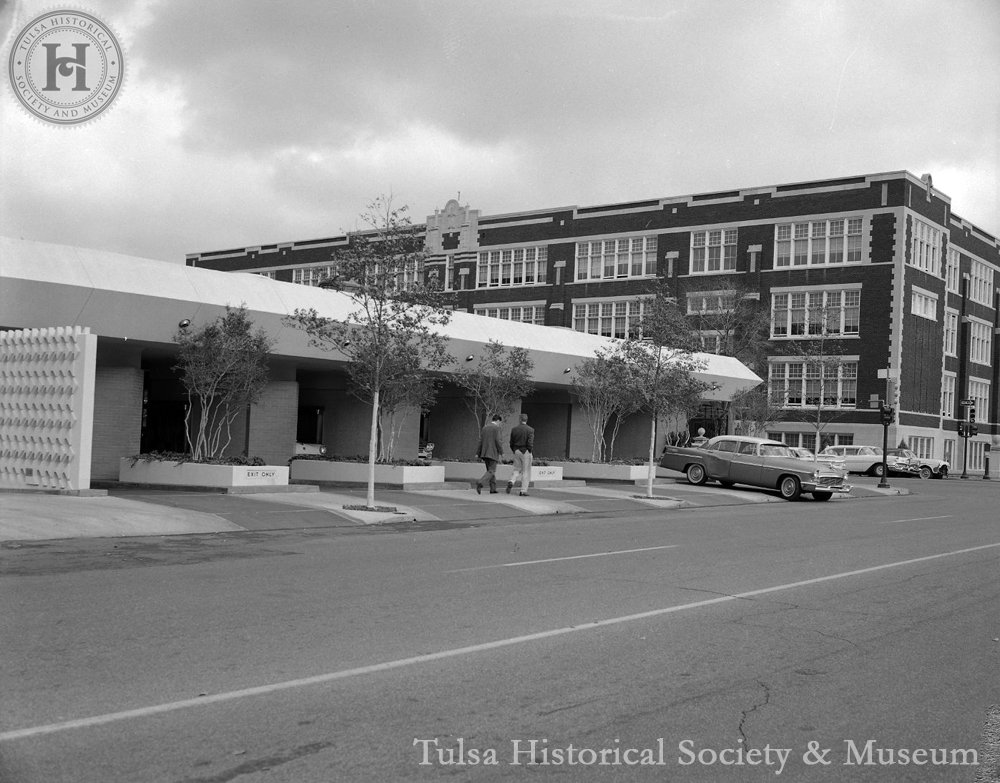 A black and white photo of a street scene featuring a modern, mid-century building with large windows, a row of small trees along the sidewalk, and a vintage car parked on the street. Two people are walking on the sidewalk.