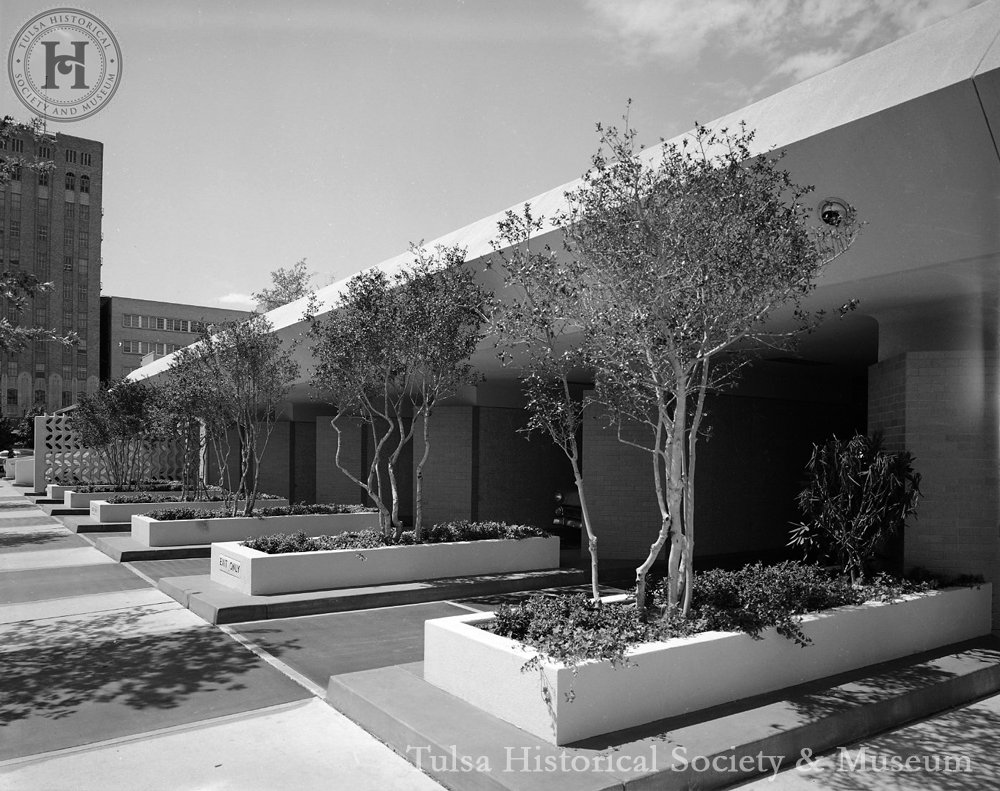 Black and white photo of a modern building exterior with rectangular planter boxes containing small trees and shrubs. A tall building is in the background under a clear sky.