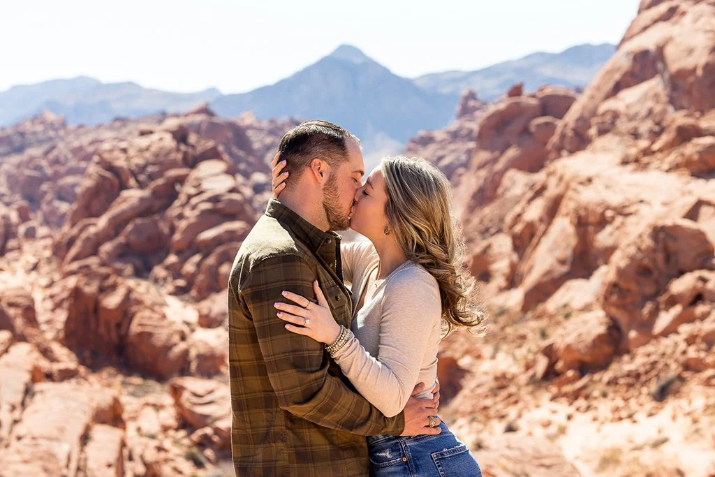 A bride and groom sharing a tender moment on their wedding day, with the groom kissing the bride's forehead, standing near a colorful floral arrangement.