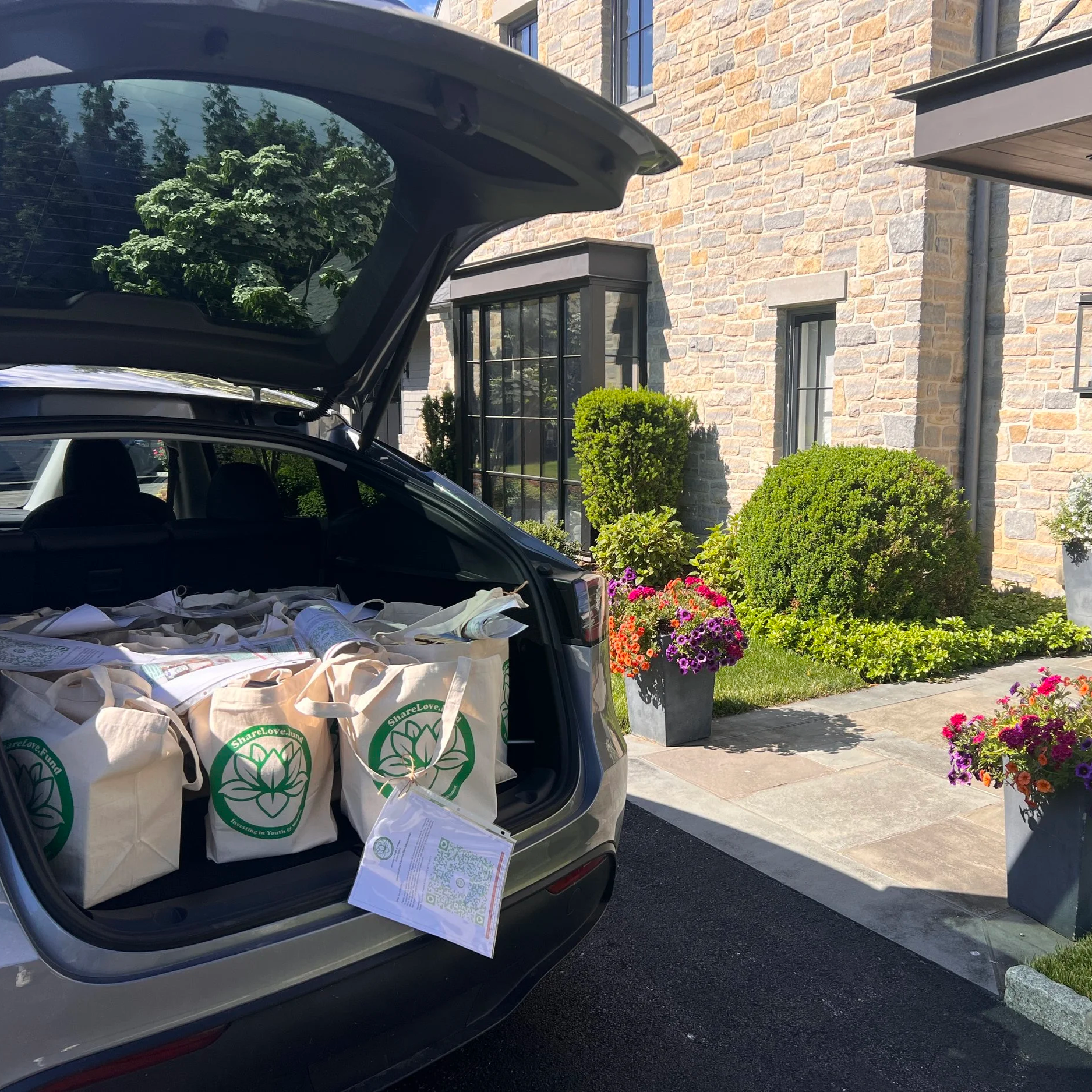 The open trunk of a gray car packed with several canvas tote bags featuring the green ShareLove.Fund lotus logo ready to be delivered by Joseluis.