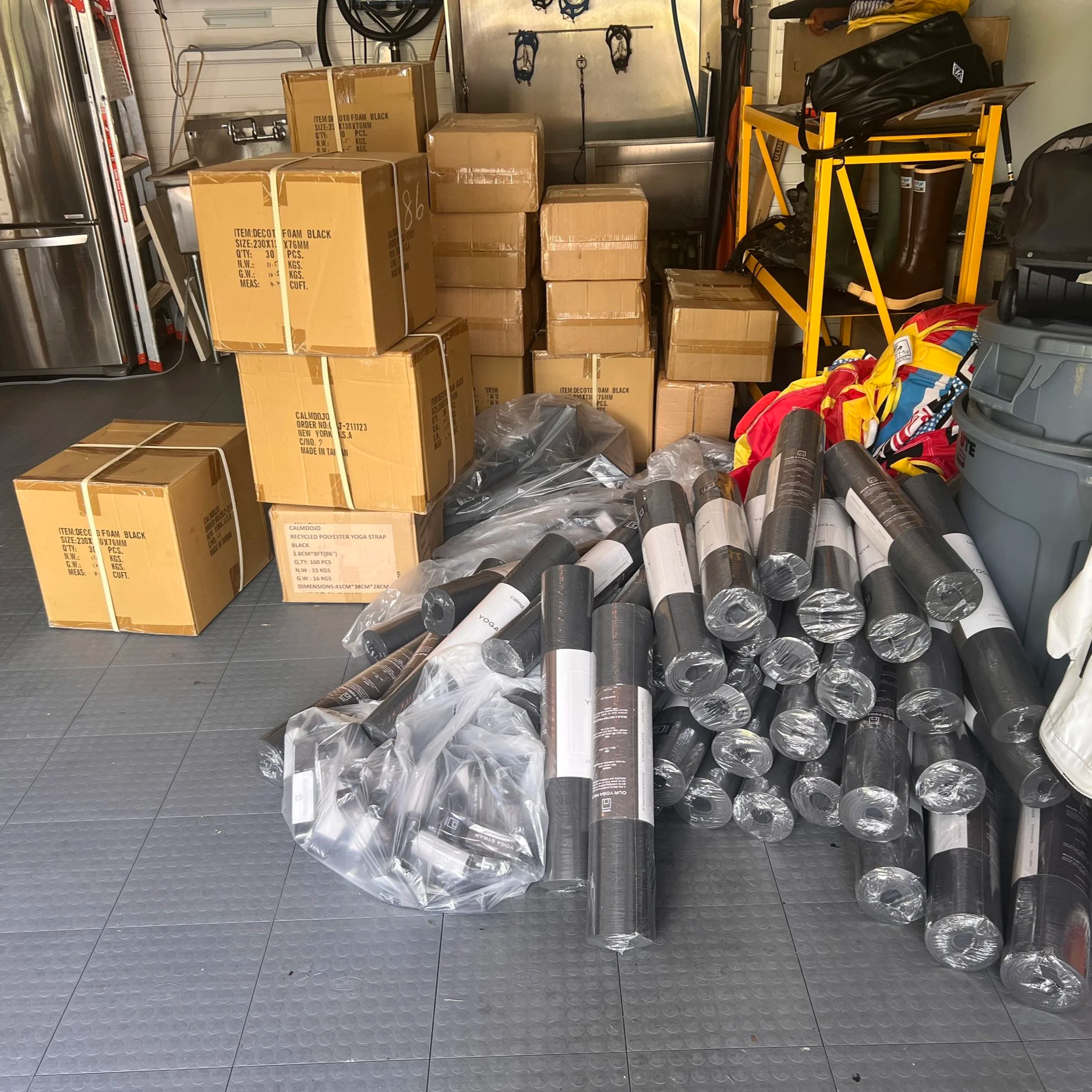 A large stack of individually wrapped, dark gray or black yoga mats rolled up on a gray tiled floor inside a storage area or garage to be donated to Rikers prison.