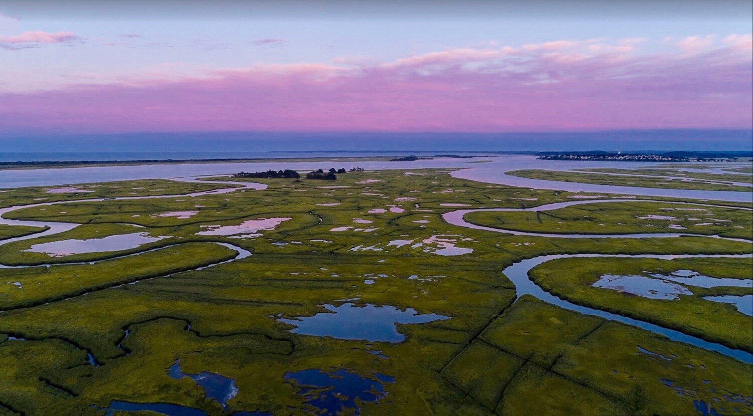 Aerial view of a marshland with winding waterways under a pink and purple sky at sunset.
