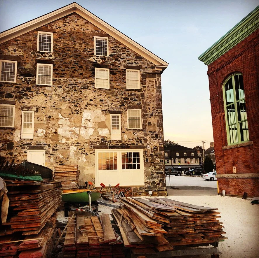 Construction site with stacks of wooden planks, a wheelbarrow, and an old stone building with multiple windows and a white door, during sunset.