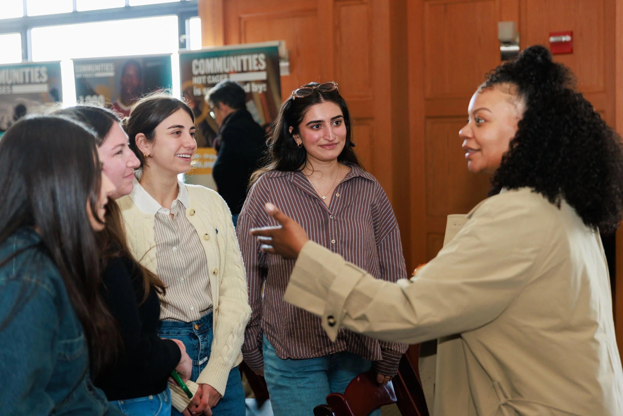 A group of women engaged in a conversation at an indoor event, with posters about communities in the background.