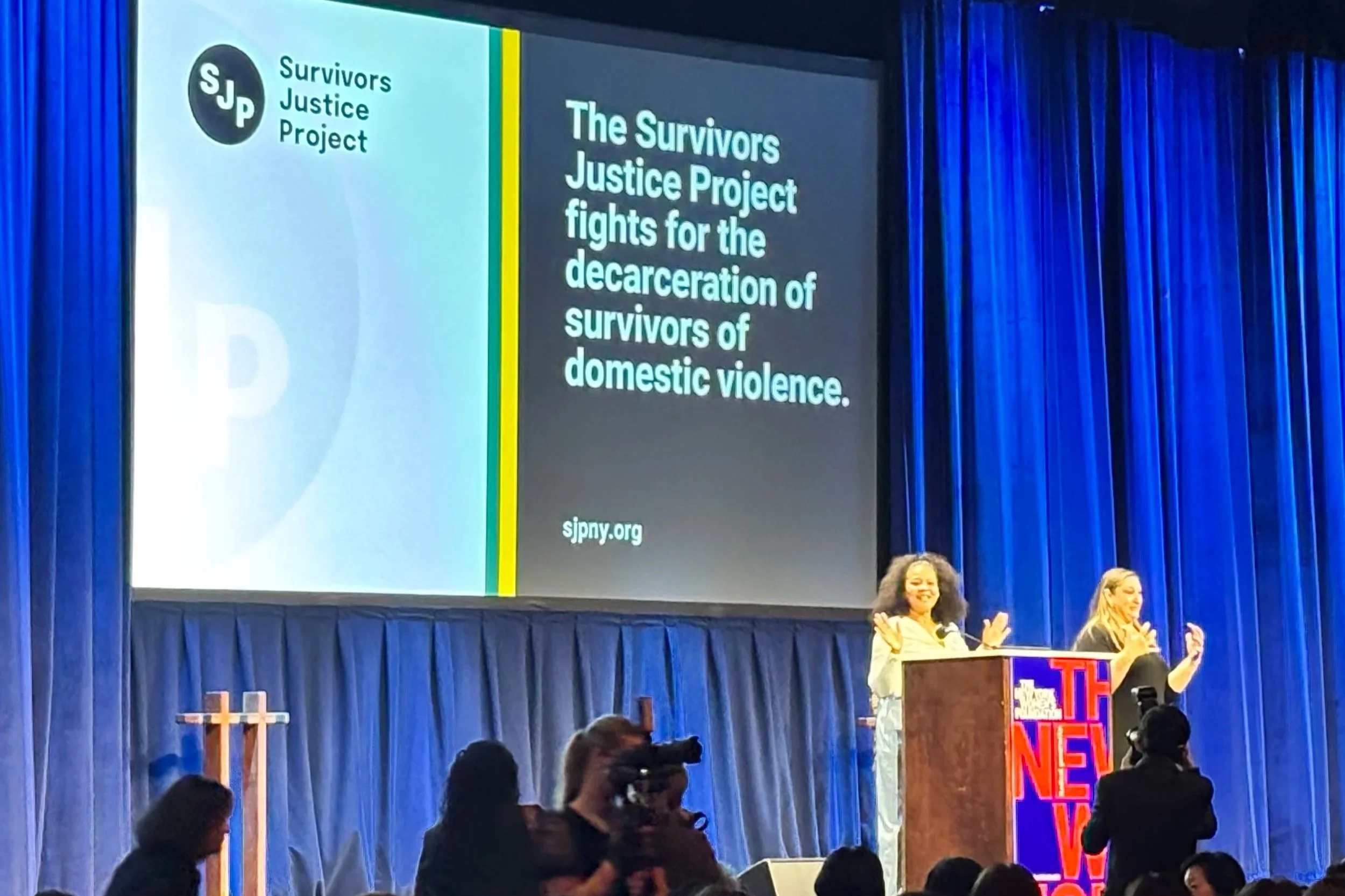A large presentation screen displays the logo and message of the Survivors Justice Project, advocating for decarceration of domestic violence survivors, with a woman speaking at a podium and an audience in a room with blue curtains.