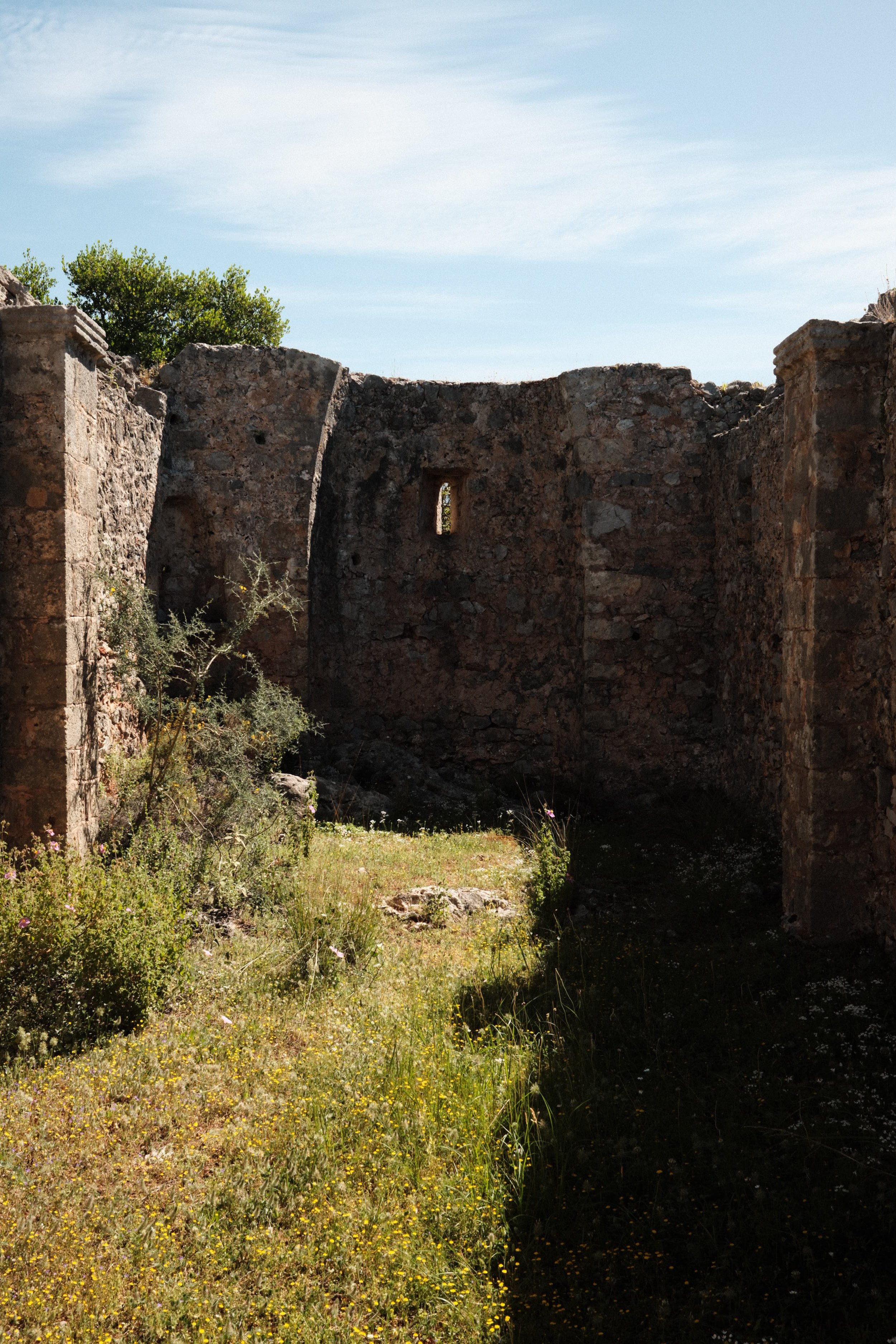 Ruins of an old stone structure with a small window, surrounded by overgrown grass and plants under a blue sky with some clouds.