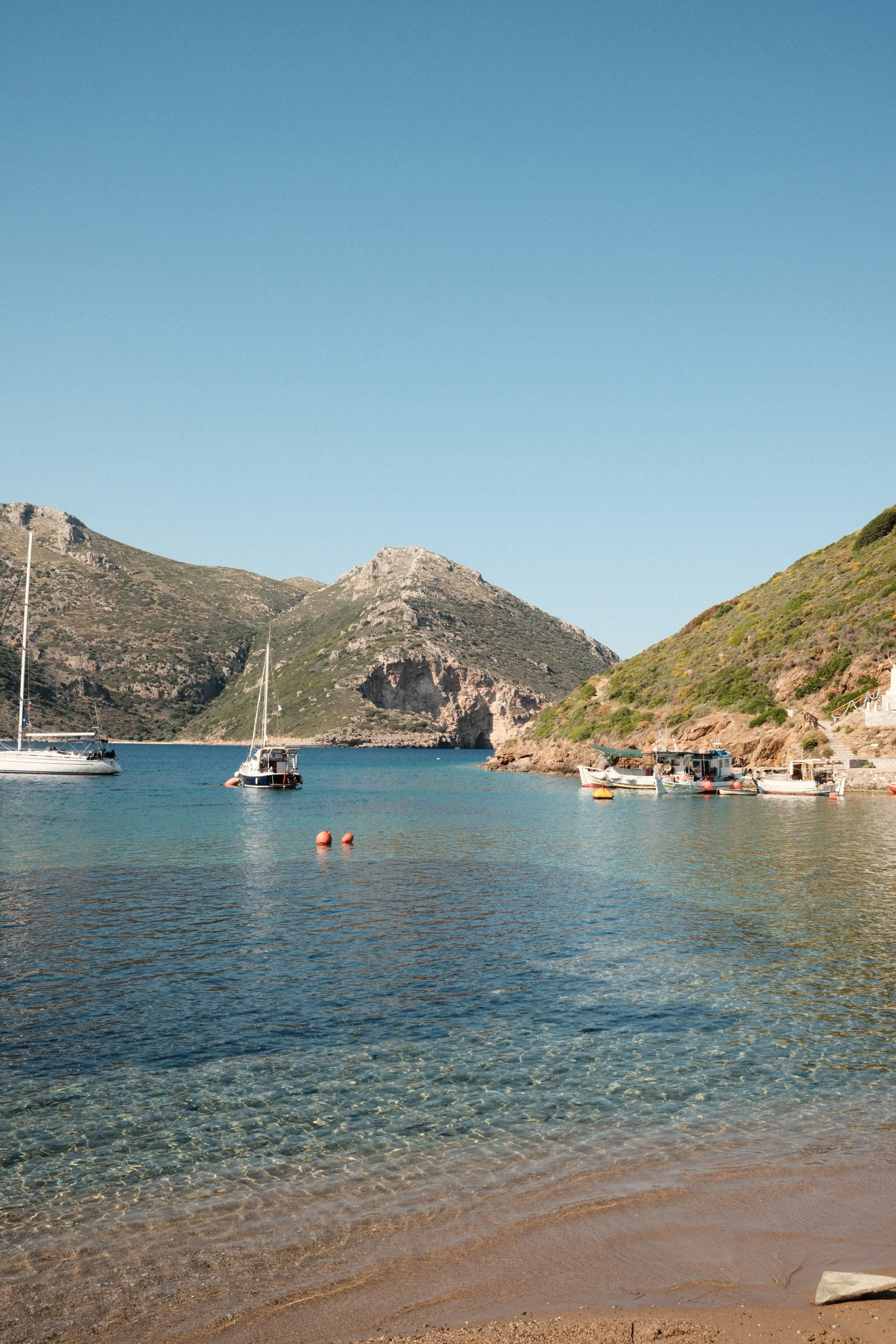 A serene coastal scene with boats anchored in calm water, surrounded by green hills and rocky cliffs under a clear blue sky.
