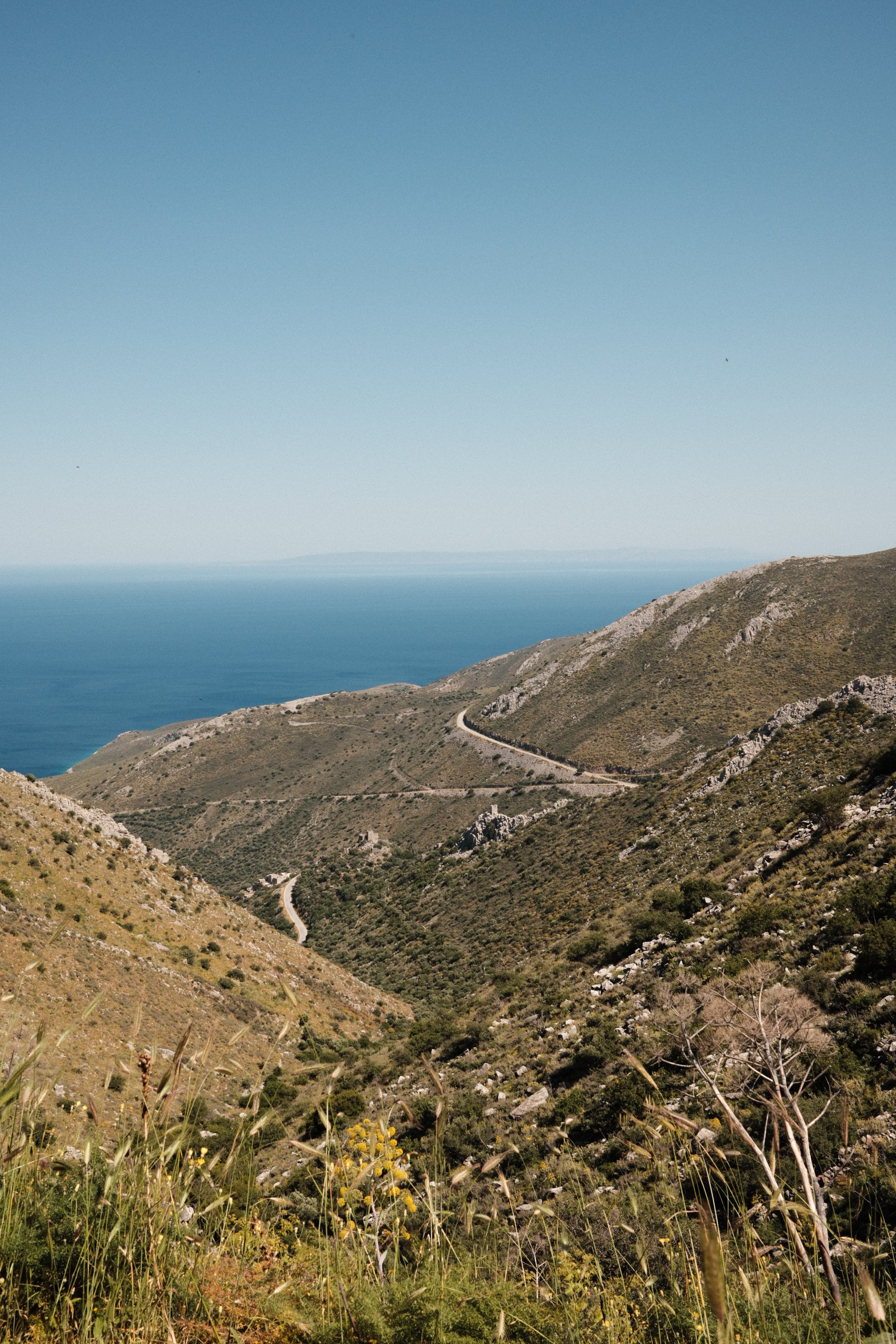 Scenic view of winding mountain road through green and rocky hills with the ocean in the distance under a clear blue sky.