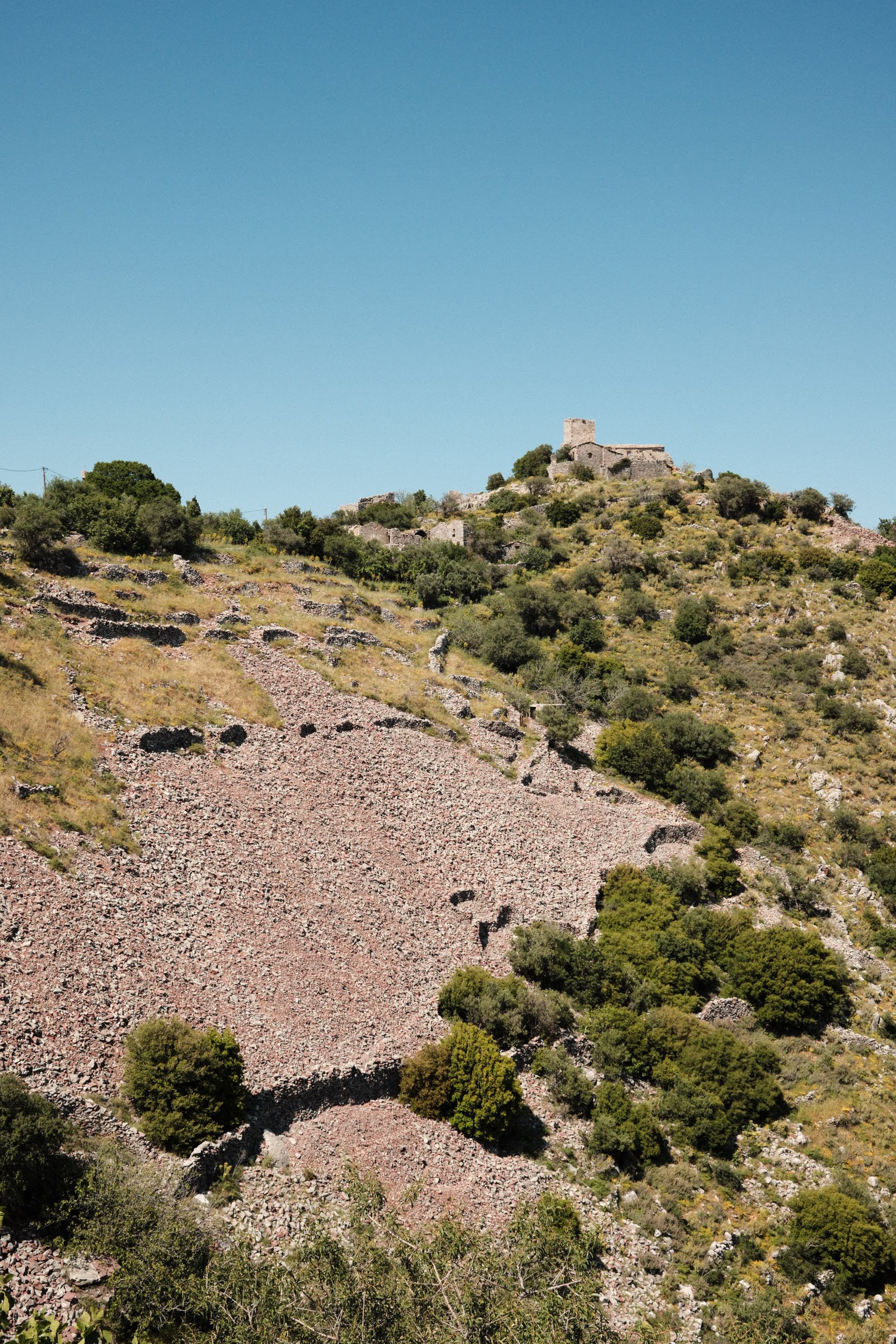 A hillside with ruins of stone structures and a small castle or tower at the top, under a clear blue sky.