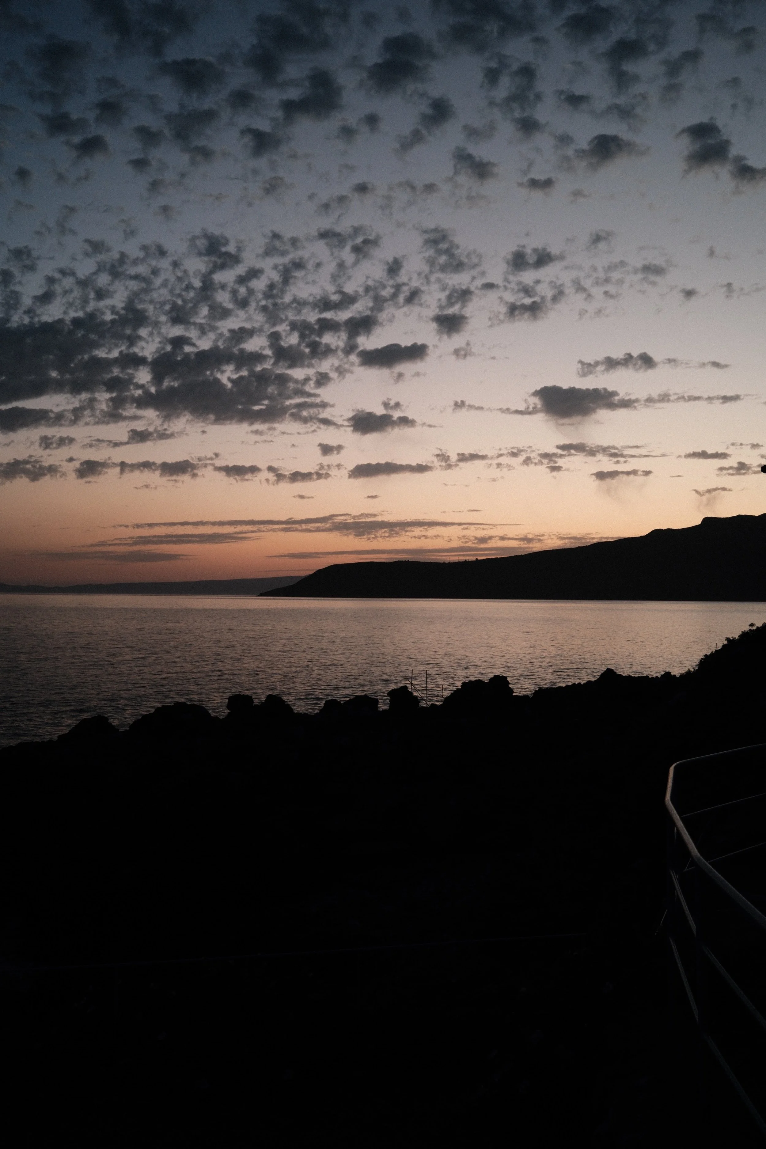 A sunset over a body of water with a silhouette of a hill or cliff in the distance and some rocks along the shoreline in the foreground.