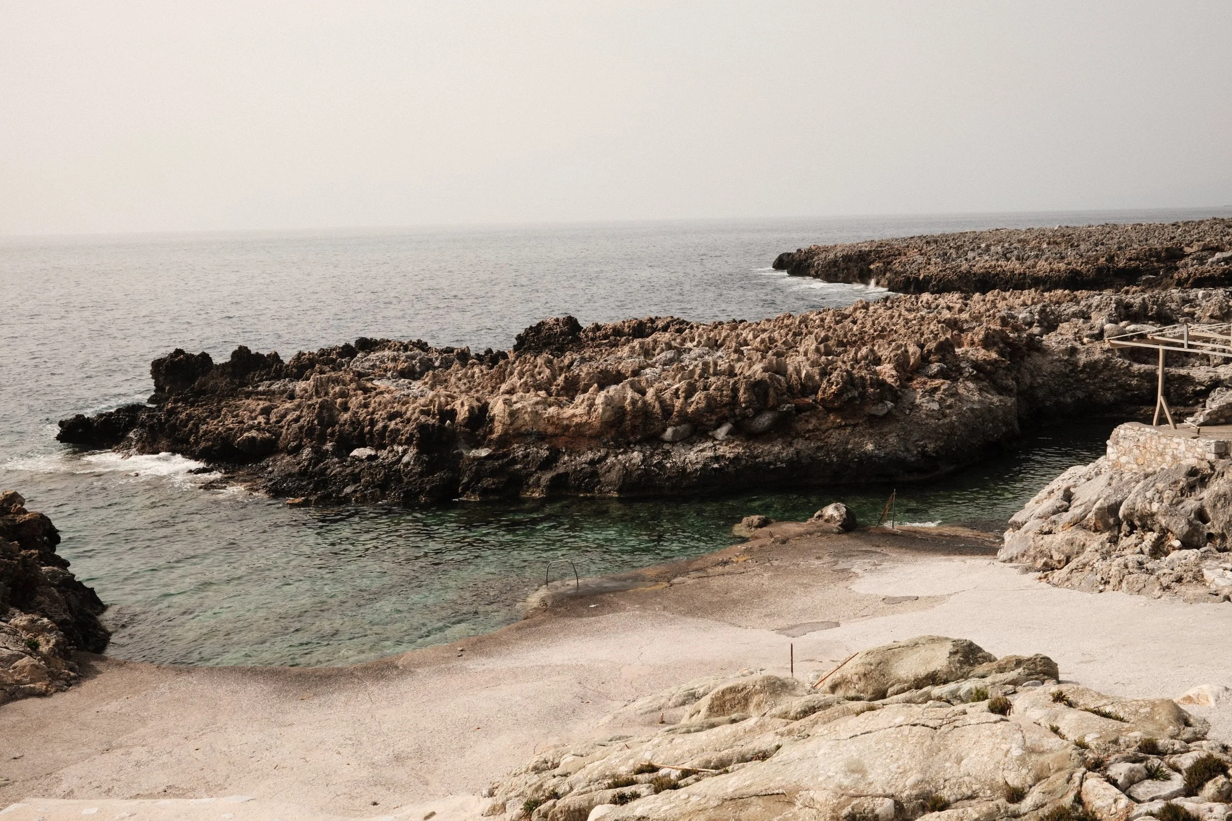Rocky coastline with a small beach and calm ocean water, overcast sky.