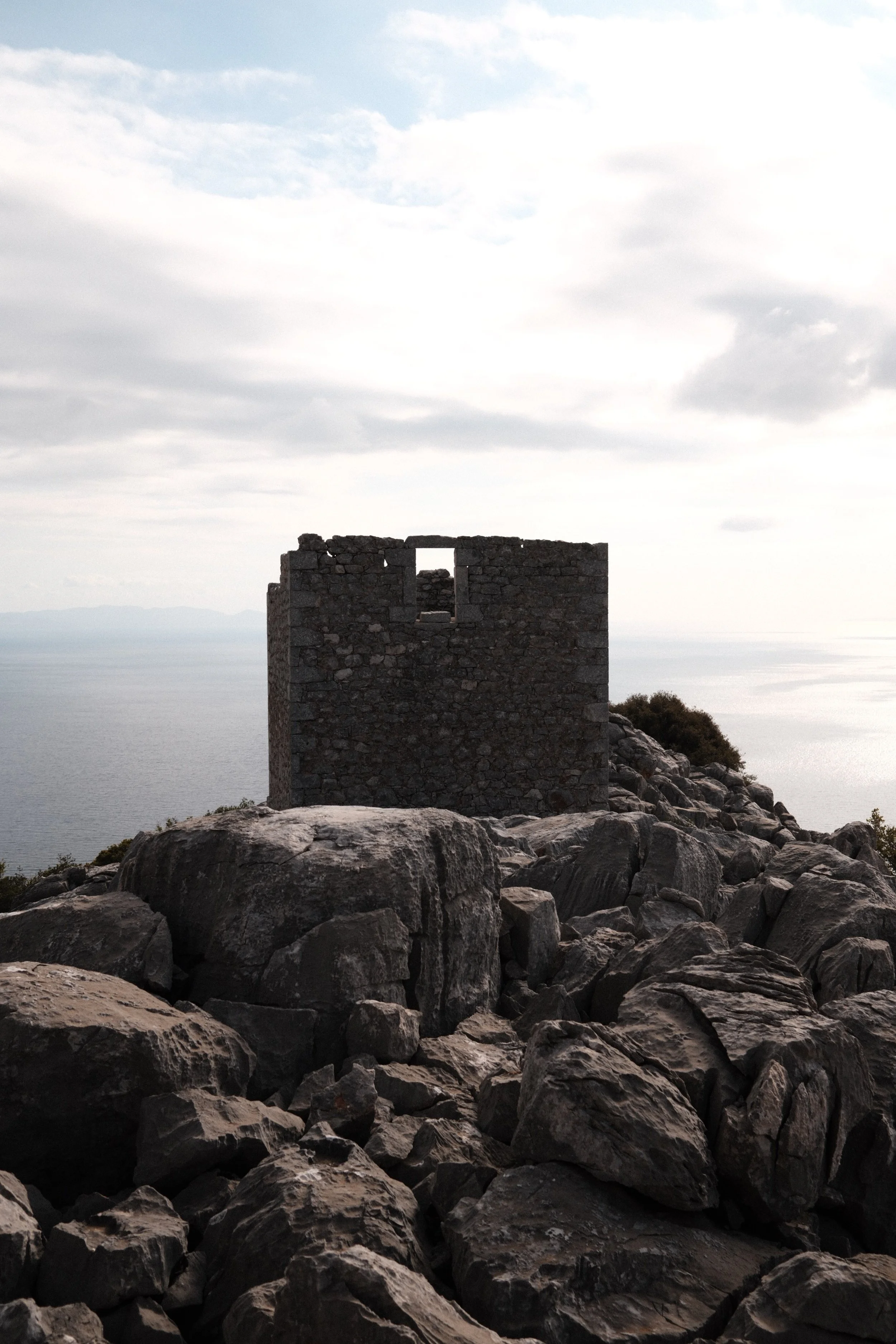 A small, abandoned stone tower on a rocky hilltop overlooking the ocean, with clouds in the sky.