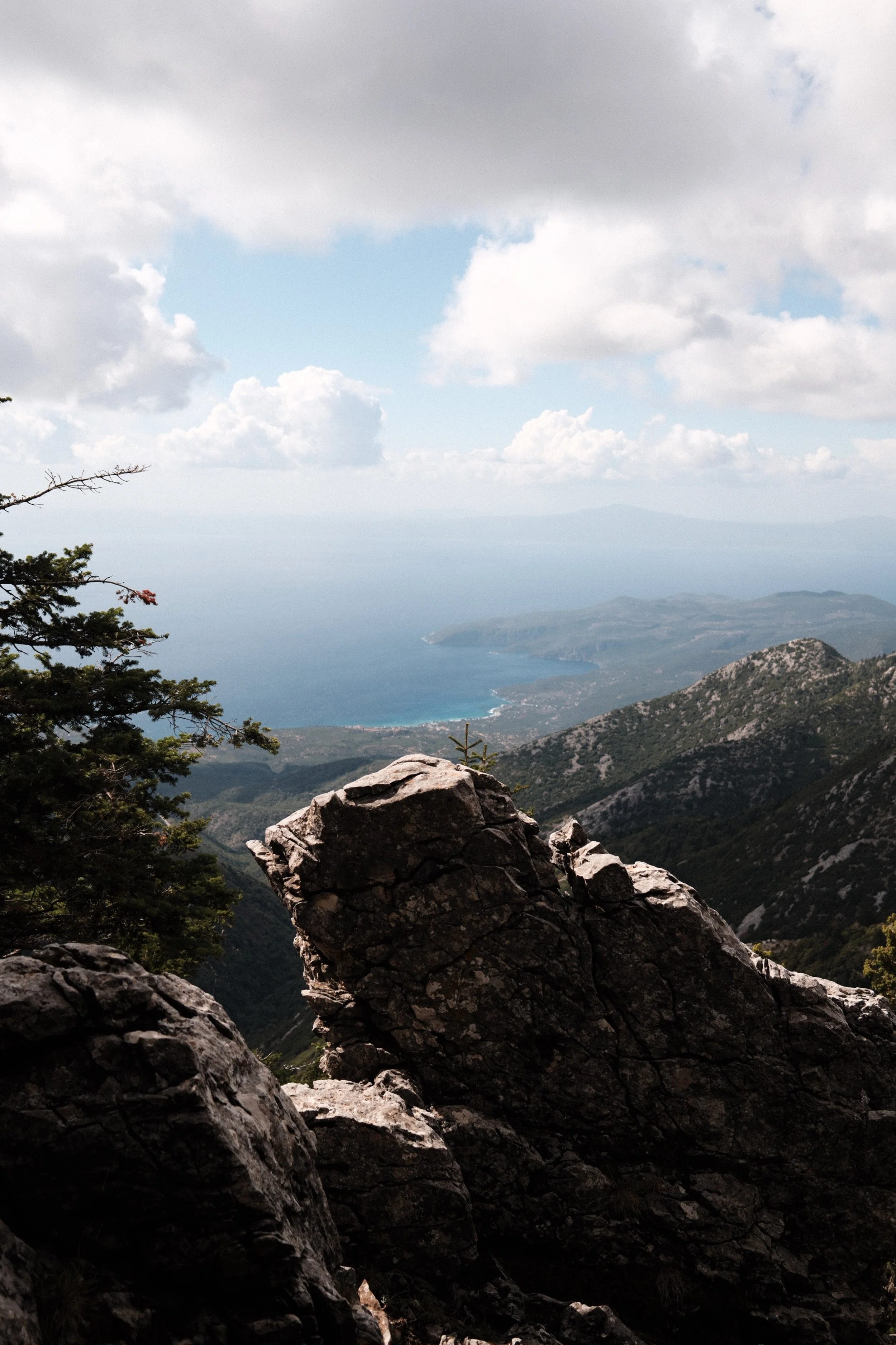 View from a mountain overlooking a coastal landscape with rocky foreground, hills, and the sea under a cloudy sky.