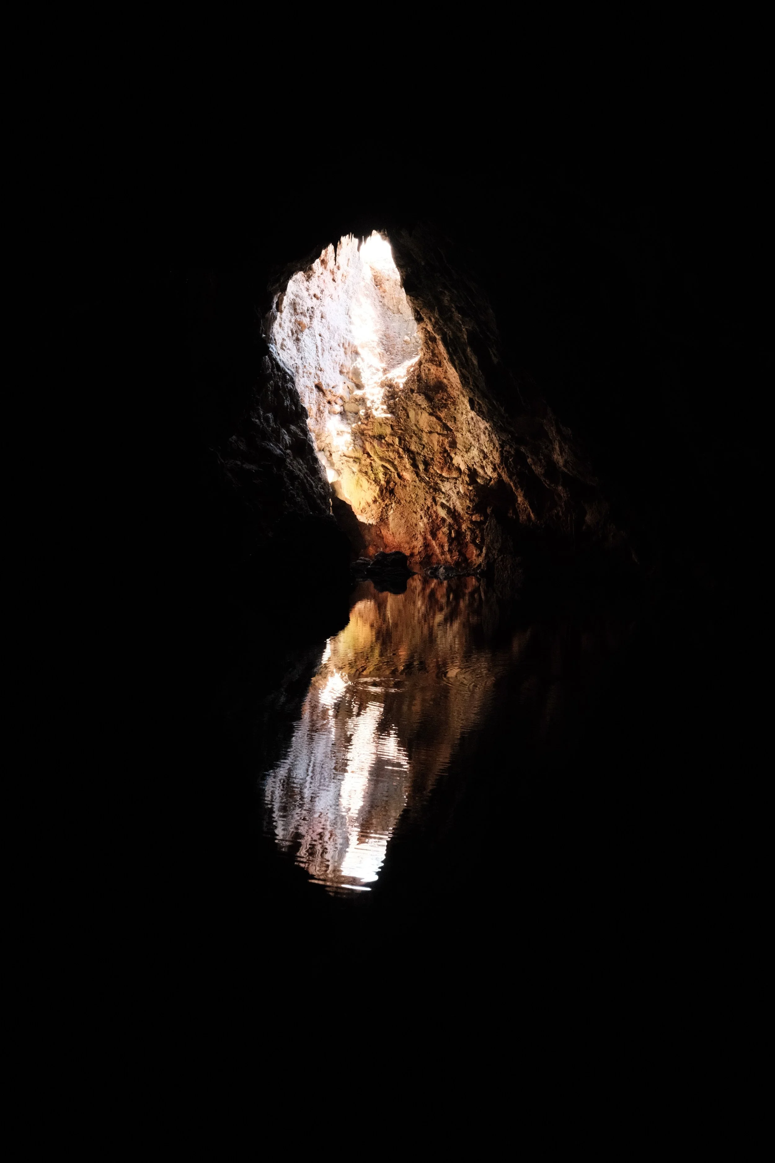 View from inside a cave looking towards the opening, with sunlight illuminating the rocky interior and reflecting on the water surface.