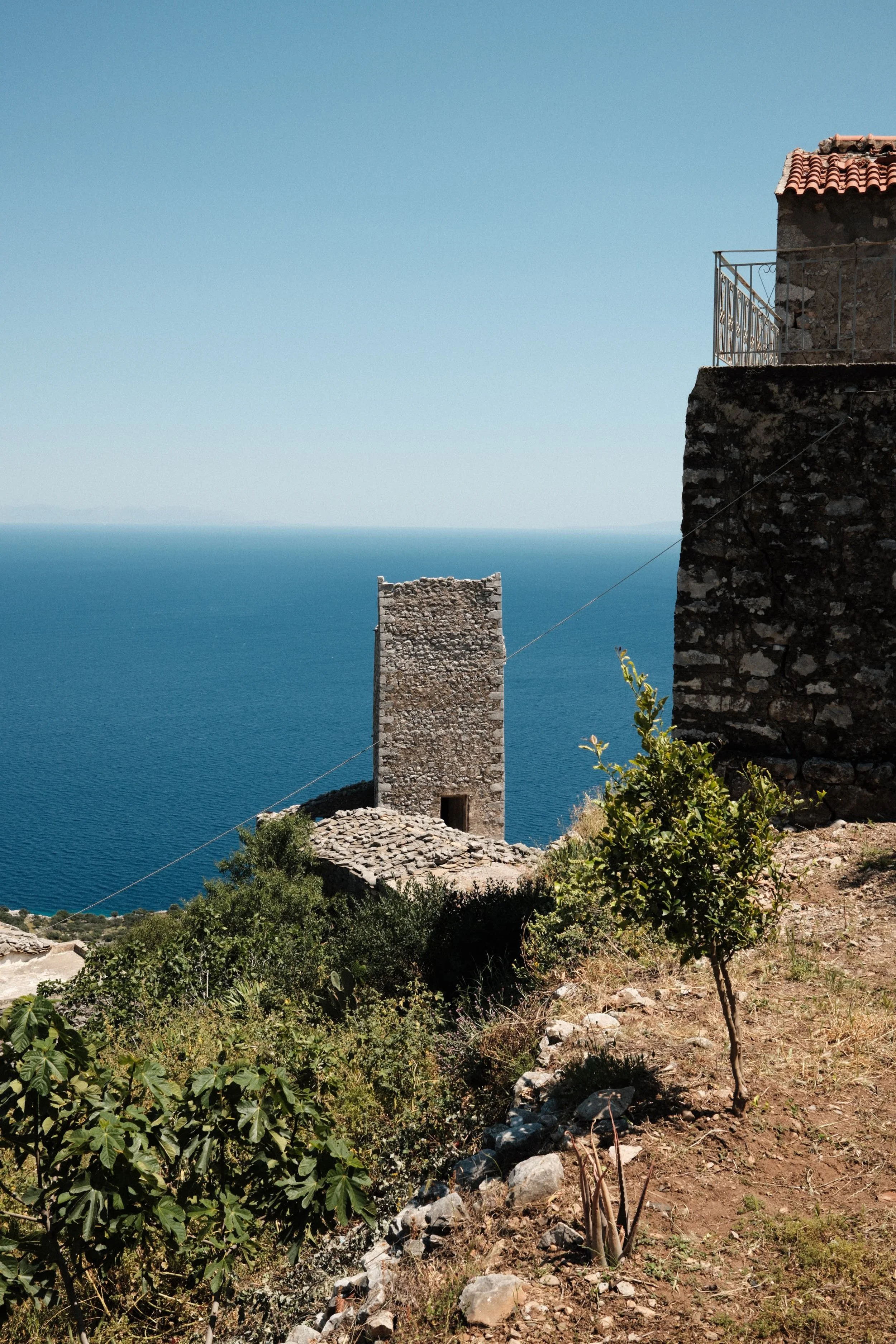 A scenic view of an ancient stone tower on a hillside overlooking the ocean, with clear blue sky above and some trees and bushes in the foreground.
