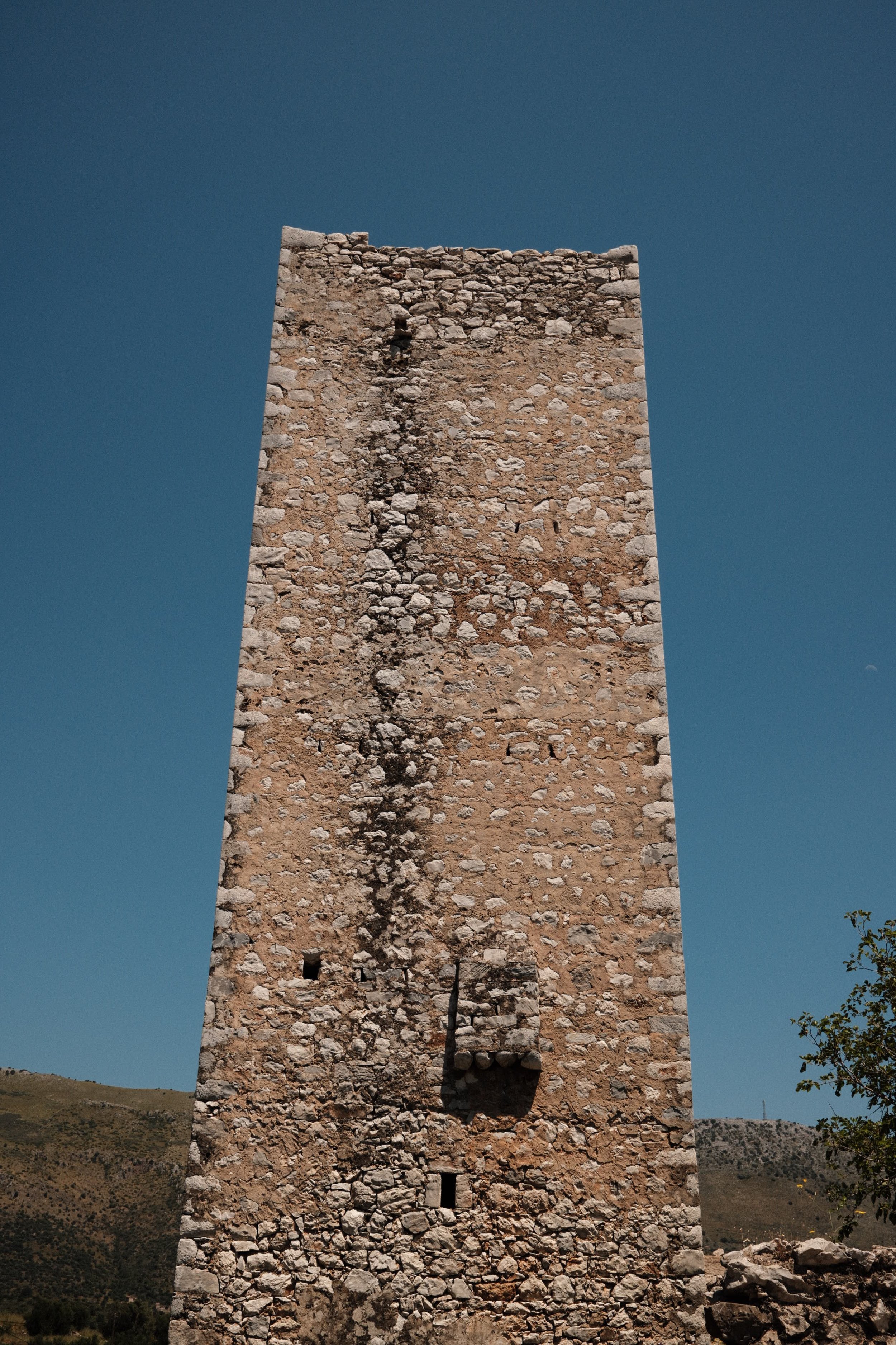 A tall, ancient stone tower with a visible crack running vertically, set against a clear blue sky with green hills in the background.