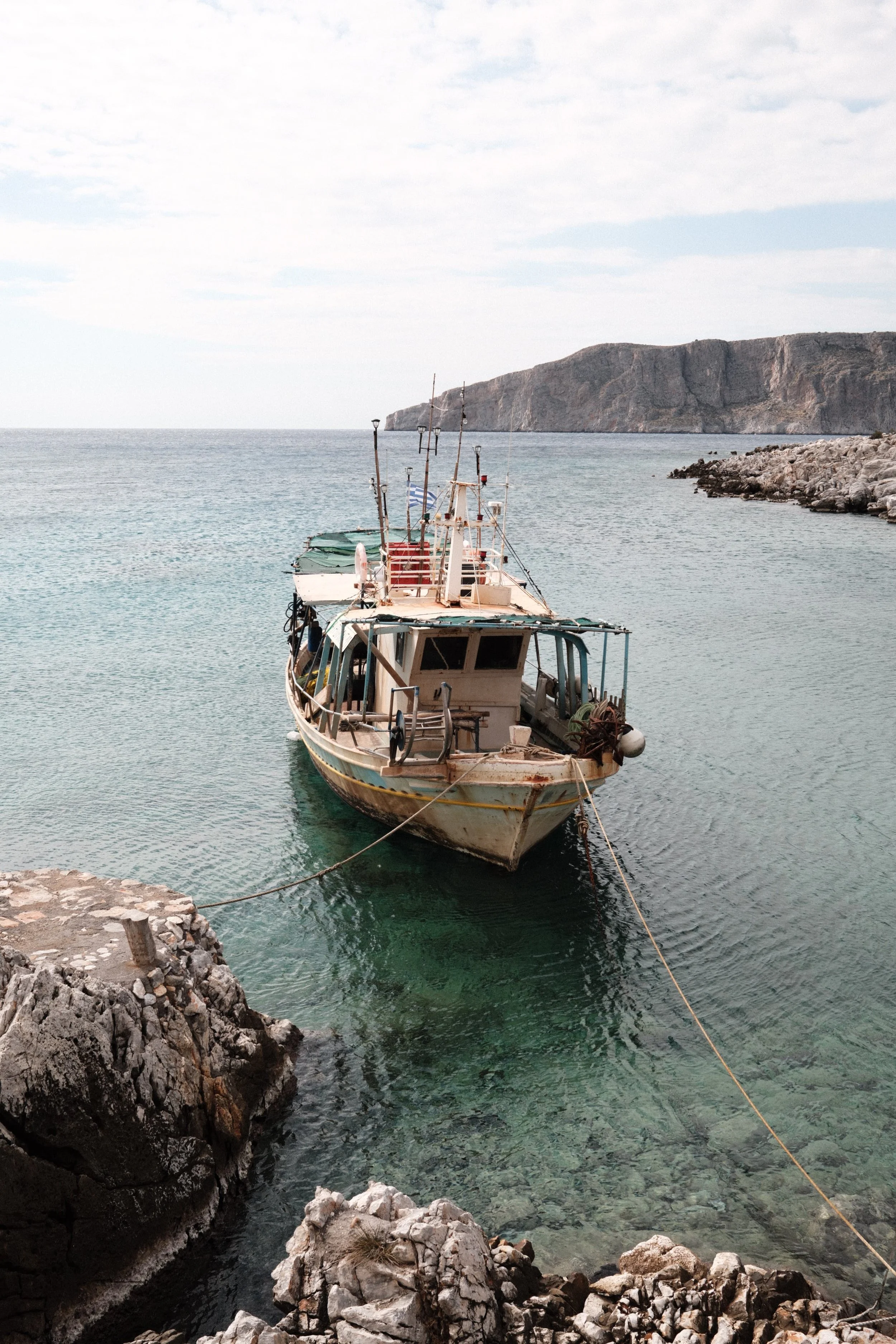 An old, rusty boat docked in clear, calm water near rocky shore, with a hillside in the background under partly cloudy sky.