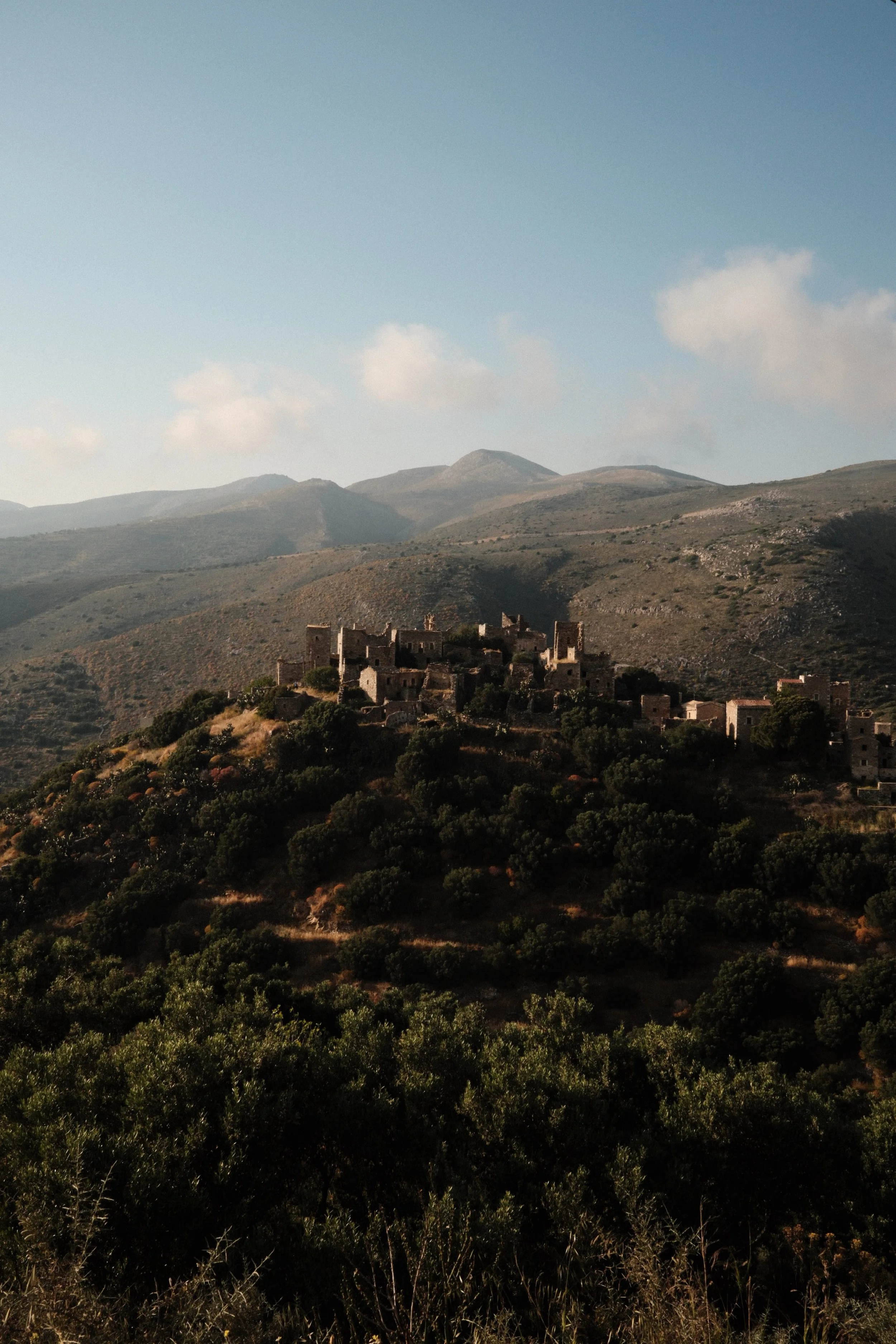 A hilltop with ancient stone ruins overlooking a landscape of rolling mountains and lush green vegetation under a partly cloudy sky.