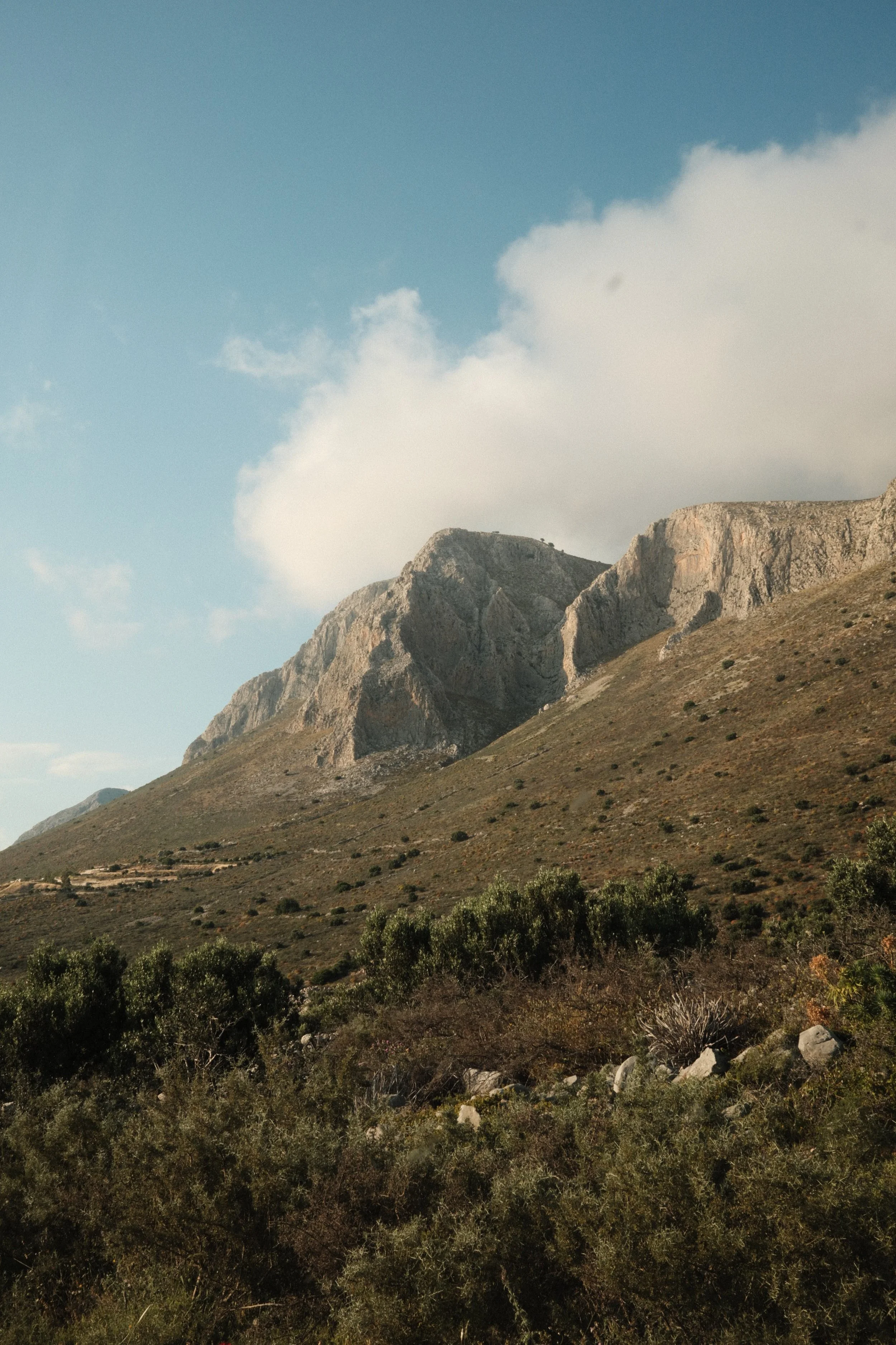 Mountain range with rugged cliffs, partly cloudy sky above, and desert vegetation in the foreground.