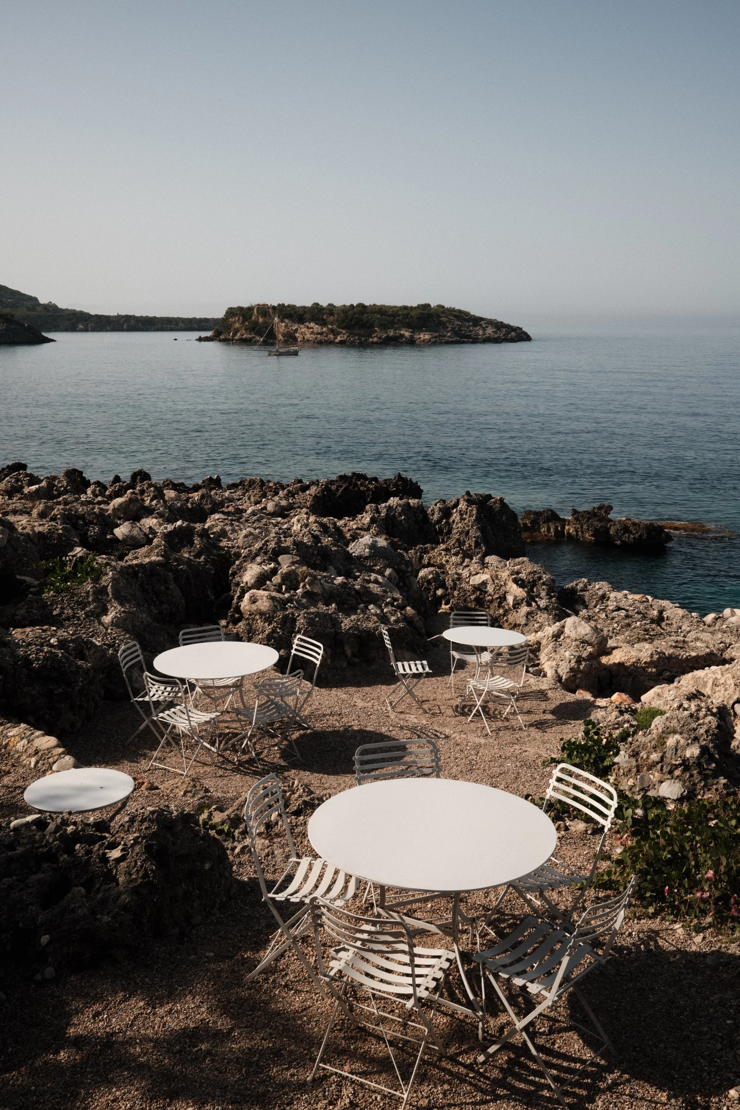 Empty white tables and chairs on rocky beach overlooking calm sea and small island with sailboat.