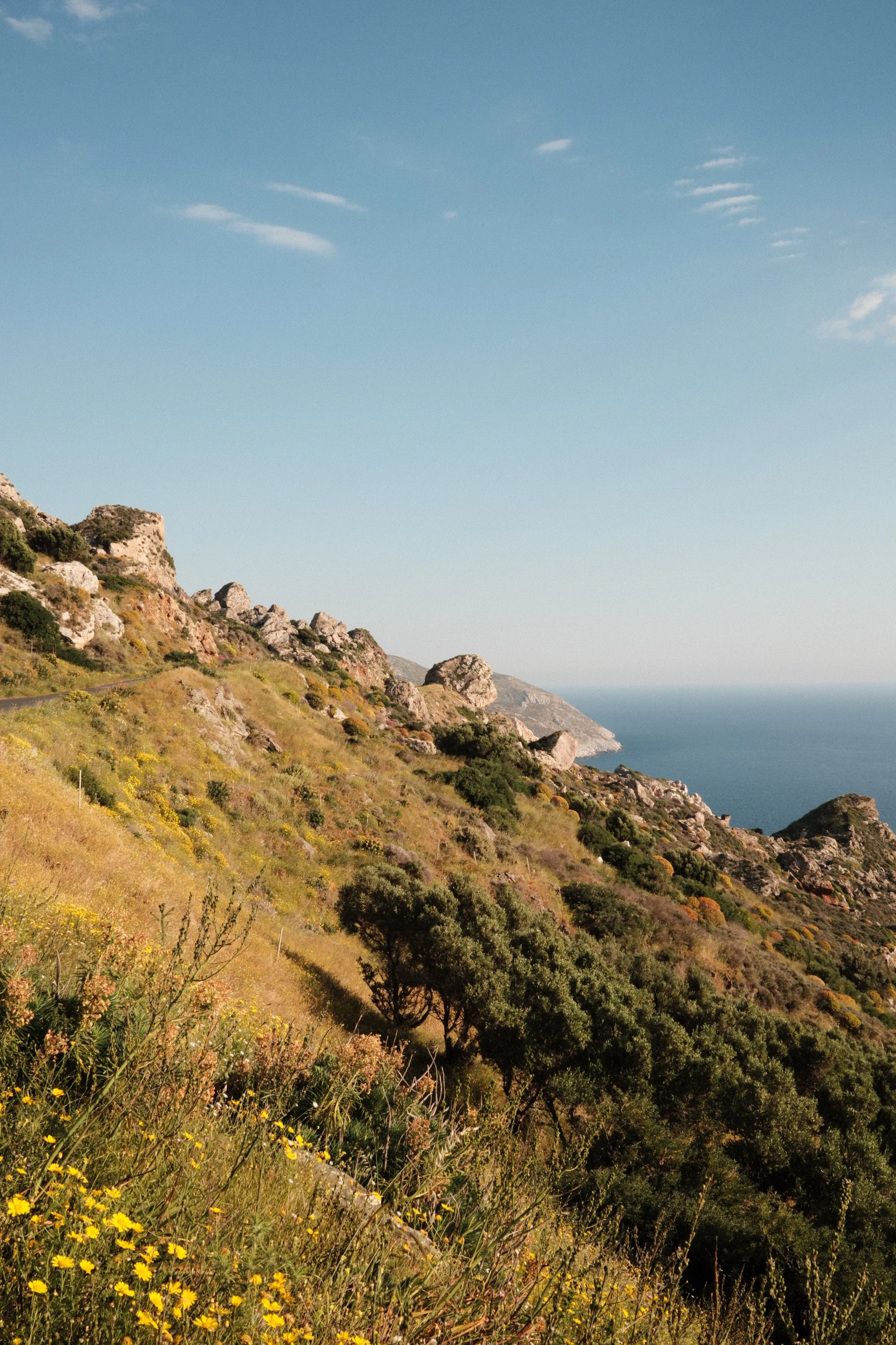 Hilly coastal landscape with rocky slopes, green and yellow vegetation, and the ocean in the background under a partly cloudy sky.