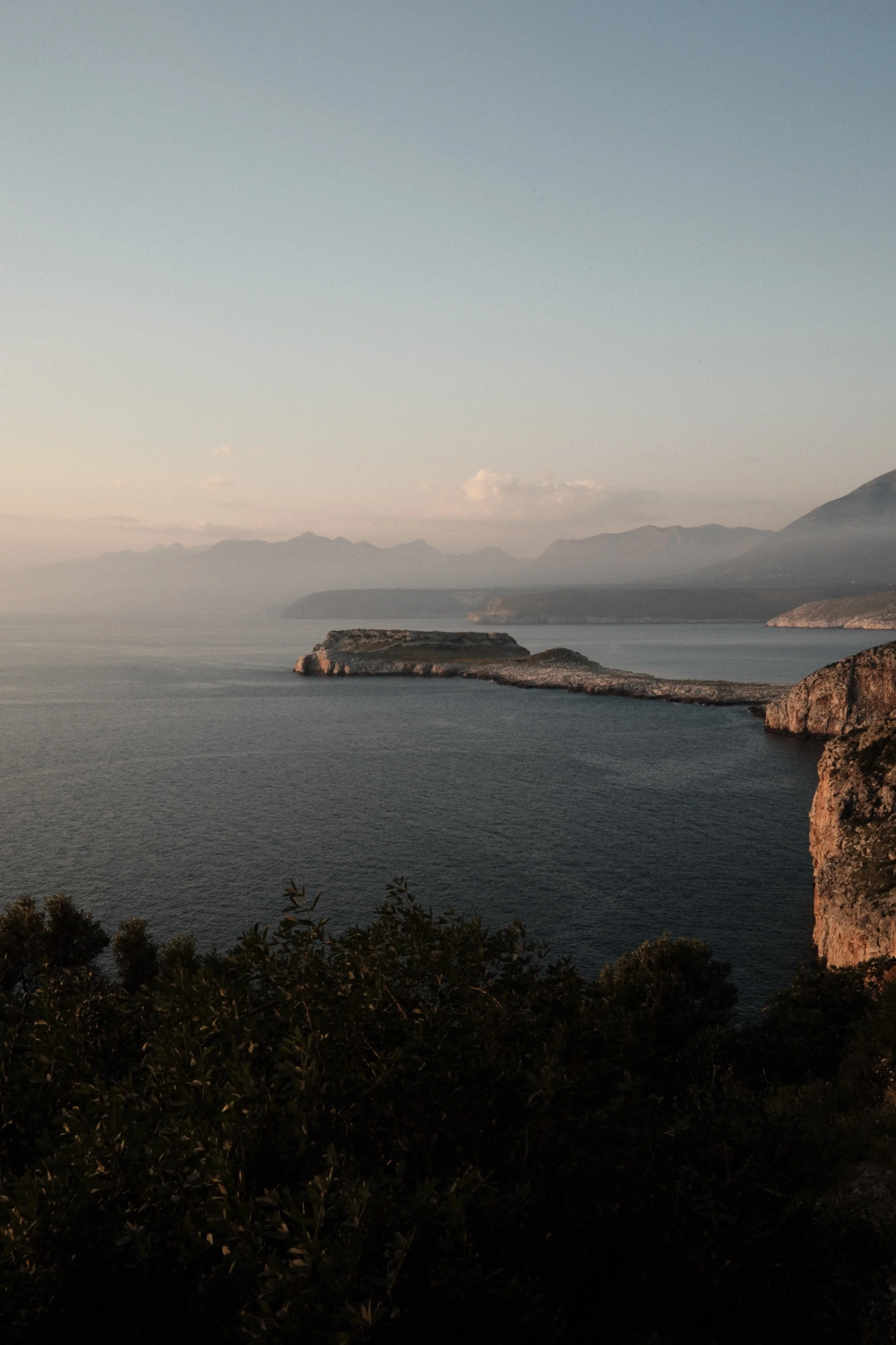 Scenic view of a calm body of water with rocky islands and mountain range in the background, under a clear sky.
