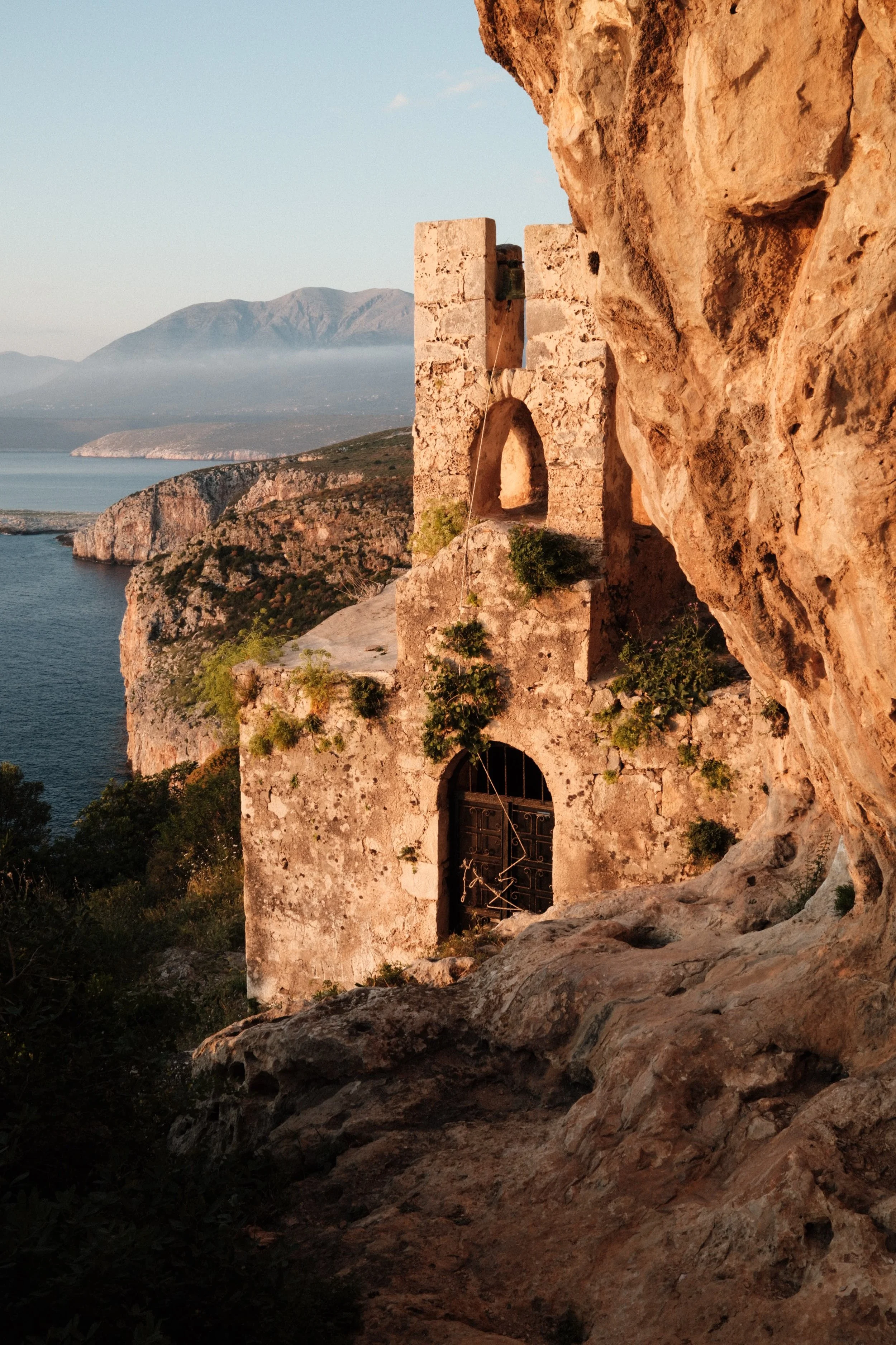 An ancient stone structure built into a rocky hillside overlooking a body of water and distant mountains, with small plants growing on its walls, during sunset.