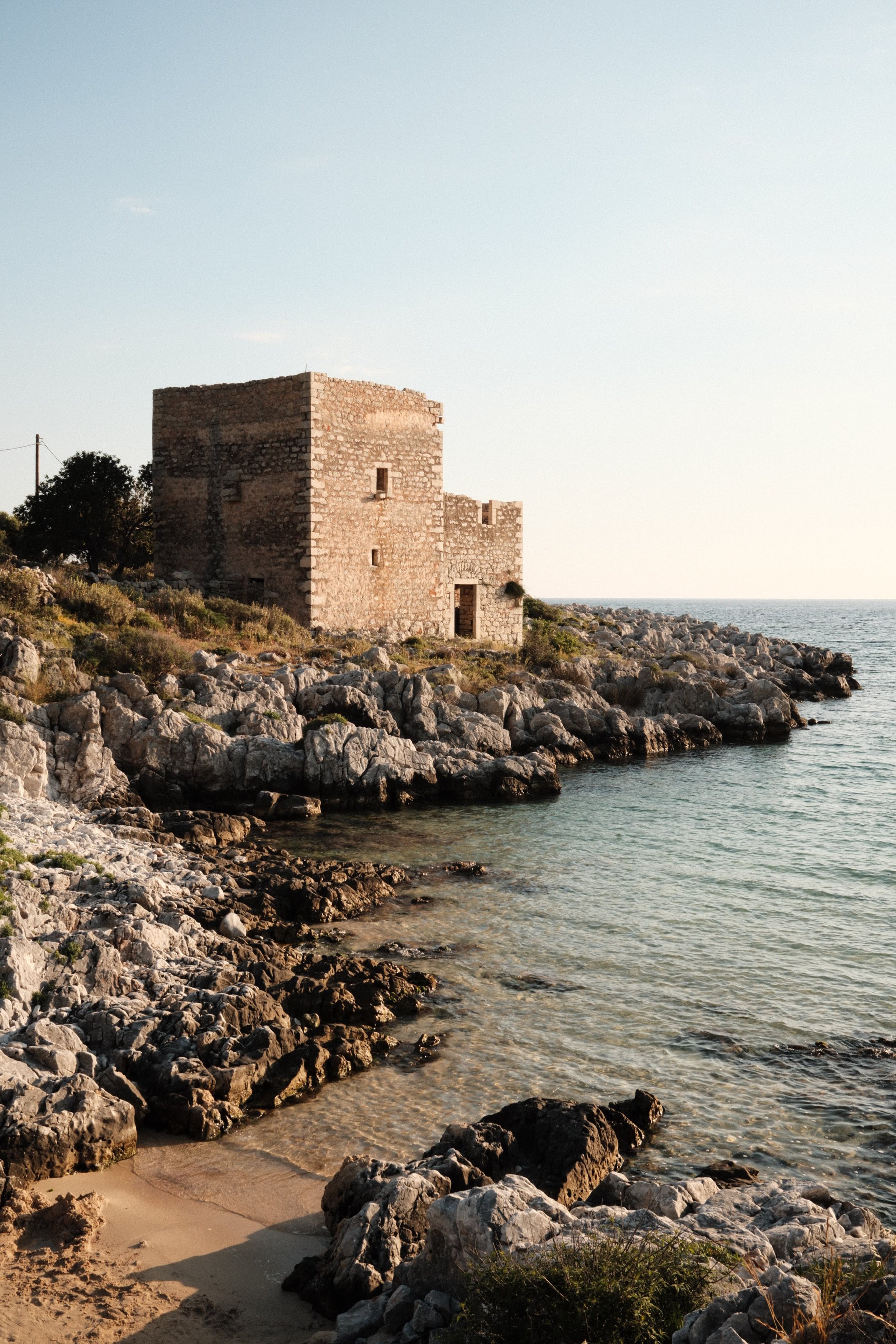 Stone castle on rocky shoreline by the sea, with calm water and clear sky during sunset or sunrise.