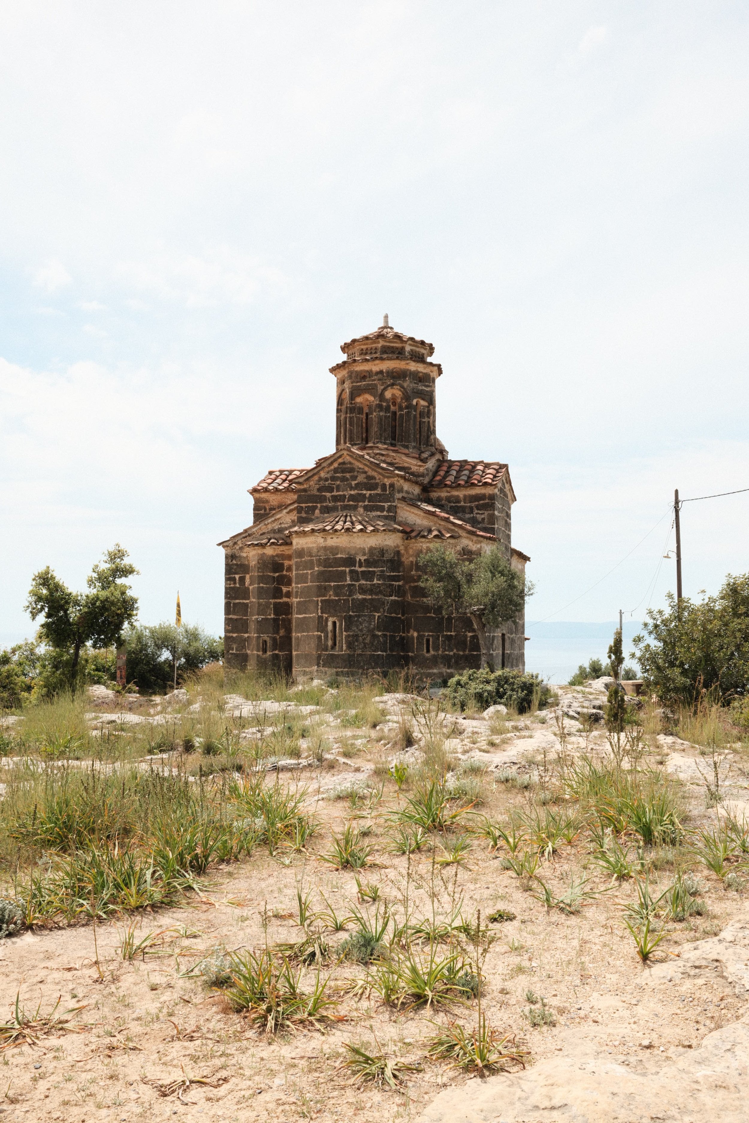 An old stone church with a red-tiled roof, situated in a sandy field with sparse grass and small bushes, under a cloudy sky.