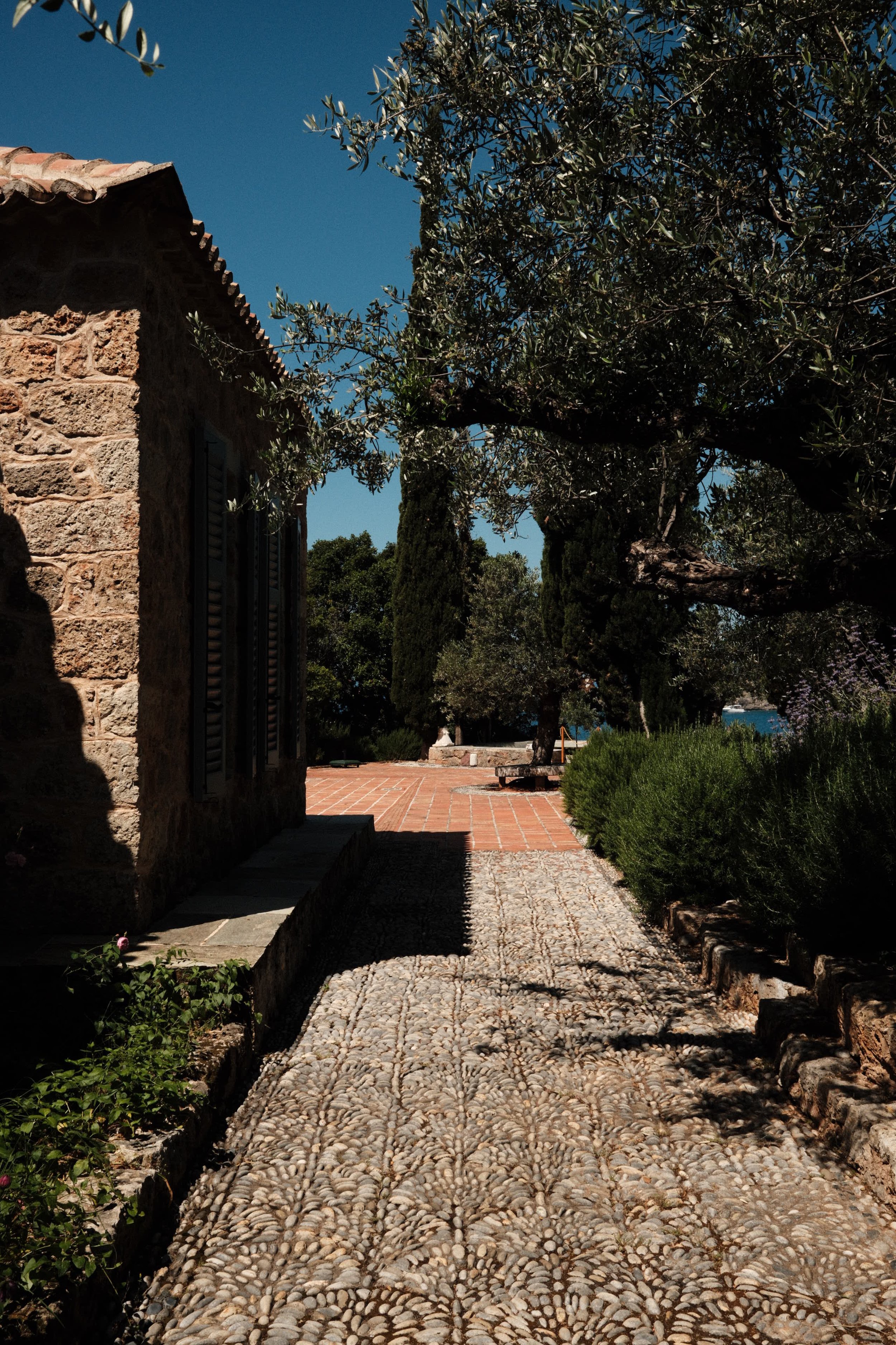 Stone pathway with pebble mosaic leading to a garden area with trees, bushes, and a stone house on the left under a clear blue sky.