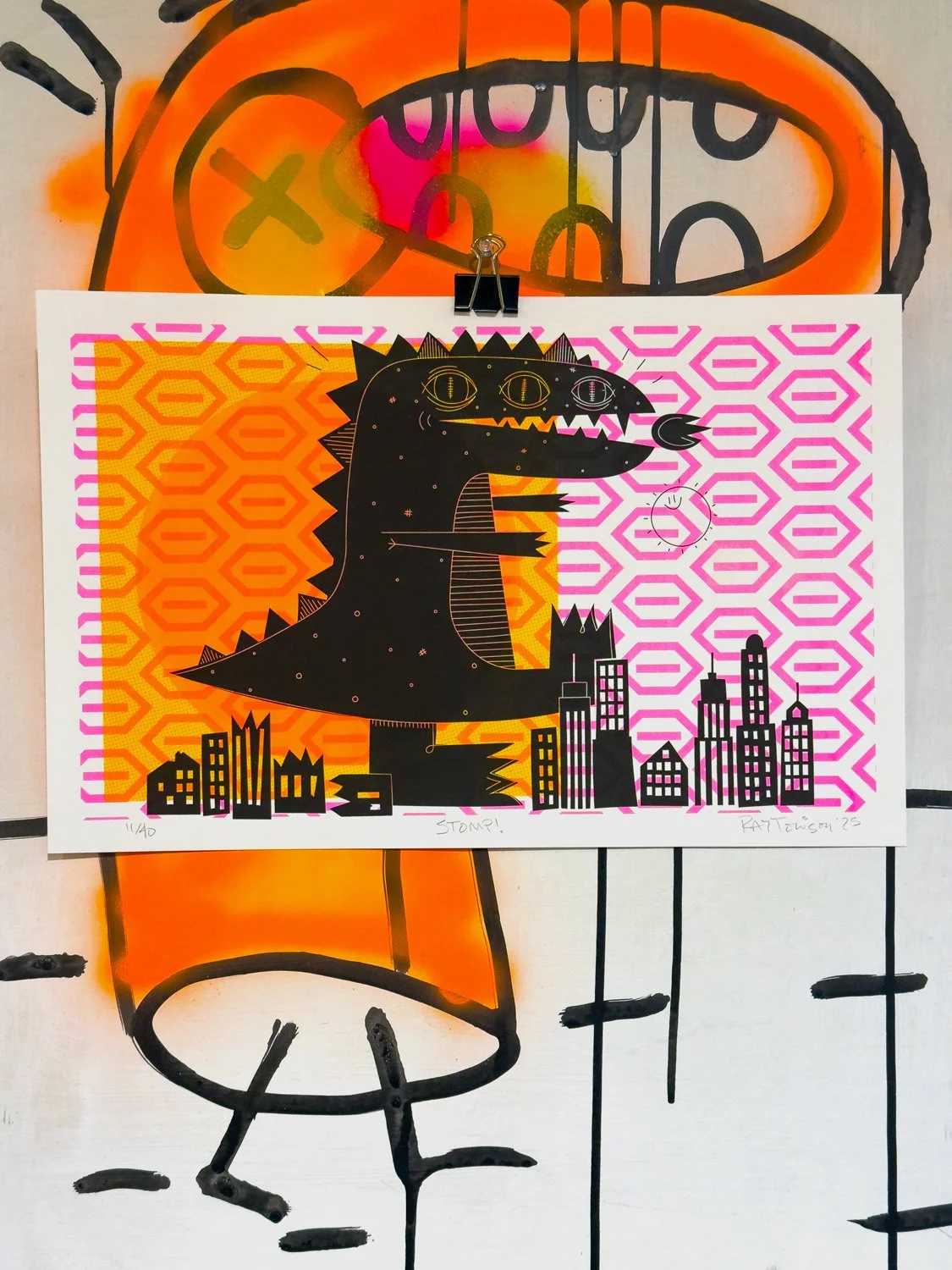 Colorful art print of a cartoon dinosaur with skyscrapers on its side, set against a pink and orange patterned background, attached to a wall with a clip.