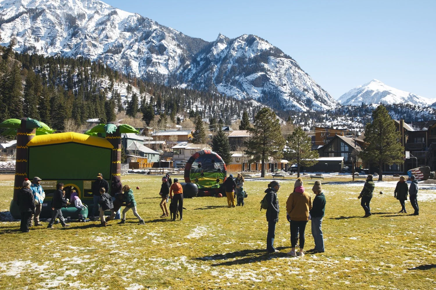 Children and adults play in bouncy houses and stand on a green field with snowy mountains in the background. 