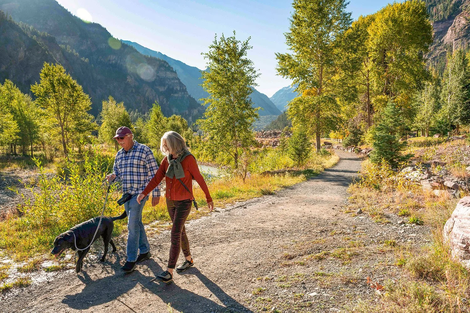 Two people and a dog walk on a flat gravel trail with trees and mountains in the background.