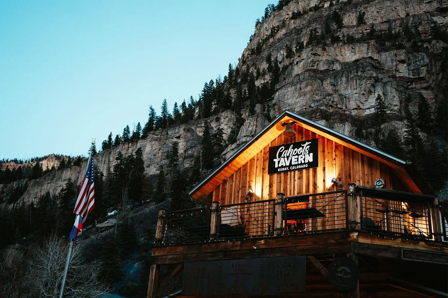 A wooden building with a porch lit up at dusk with an american flag nearby and cliffs in the background.