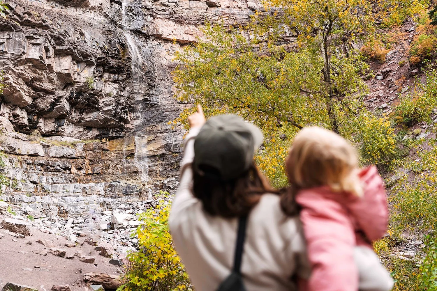 A woman points at a waterfall while holding a toddler.