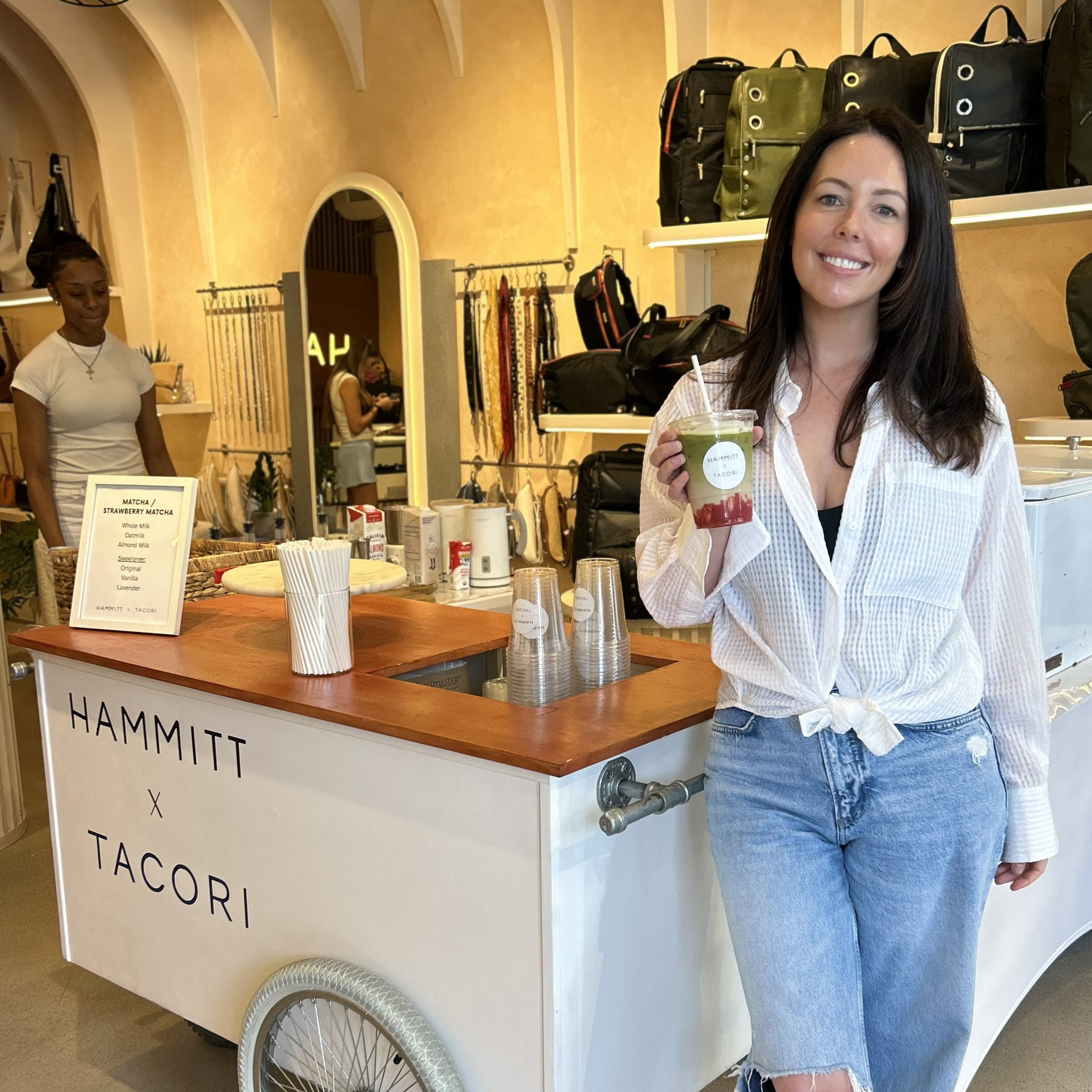 a woman standing next to a matcha cart with Hammitt and Tacori custom branding and holding a matcha drink in hand with the same custom logo