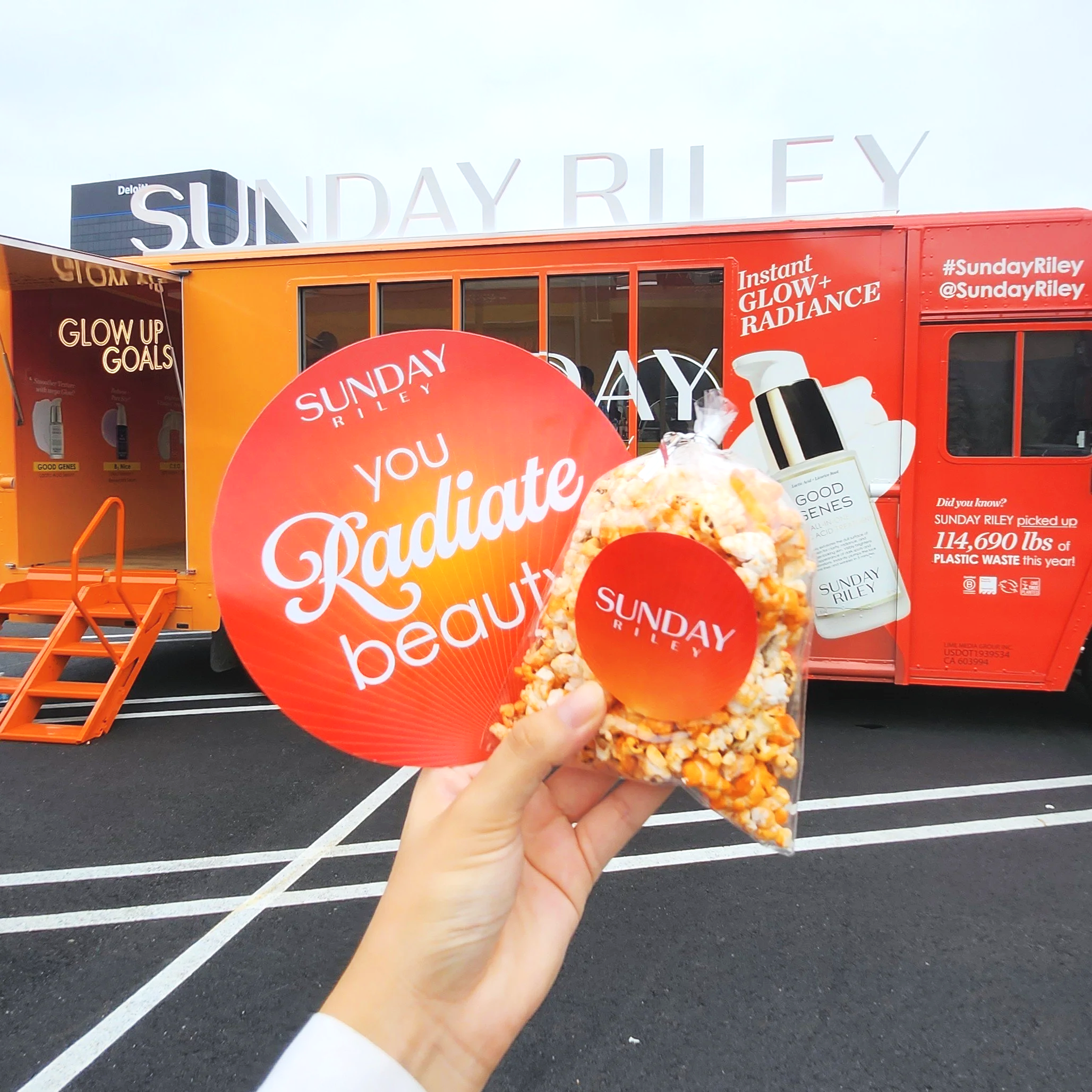 A bag of orange colored popcorn with a Sunday Riley logo in front of an orange Sunday Riley tour bus