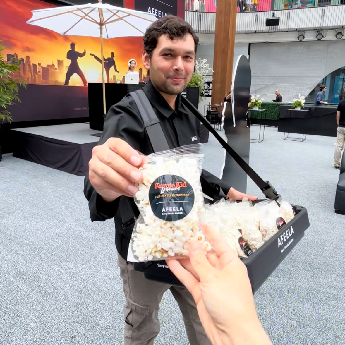 A man carrying a tray full of packaged popcorn hands one bag with label to someone