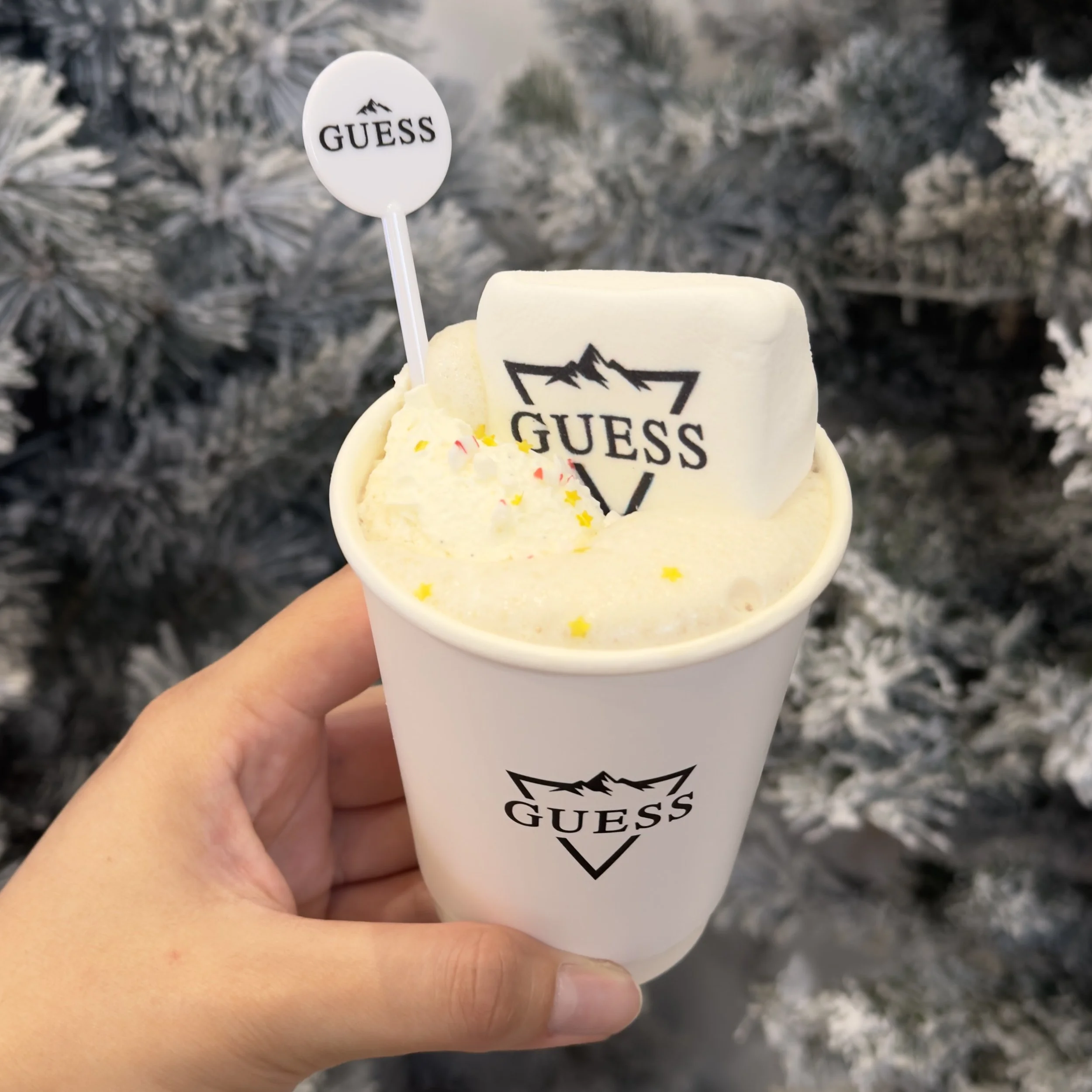 A close up of a hot chocolate cup at a brand holiday event with an imprinted Guess Jeans logo on the cup, on a marshmallow and on a drink mixer, topped with whipped cream and edible glitter