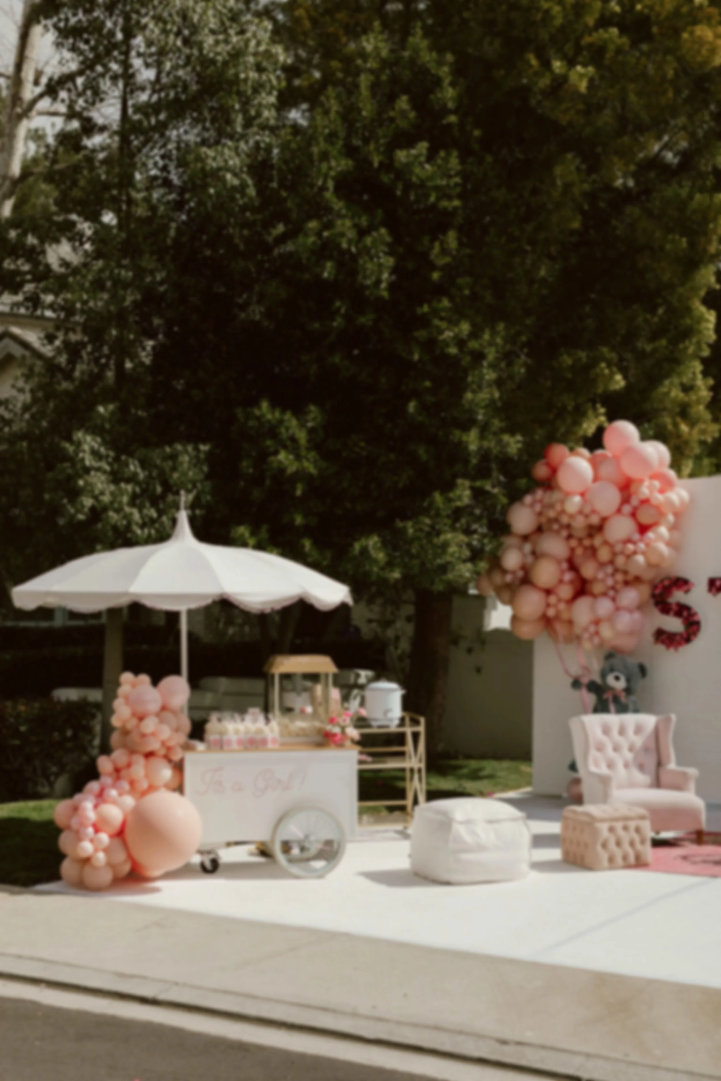 A popcorn cart for a baby shower perfectly set beneath a wooded canopy and beautifully adorned with pastel balloons, white umbrella, florals, and popcorn favors.