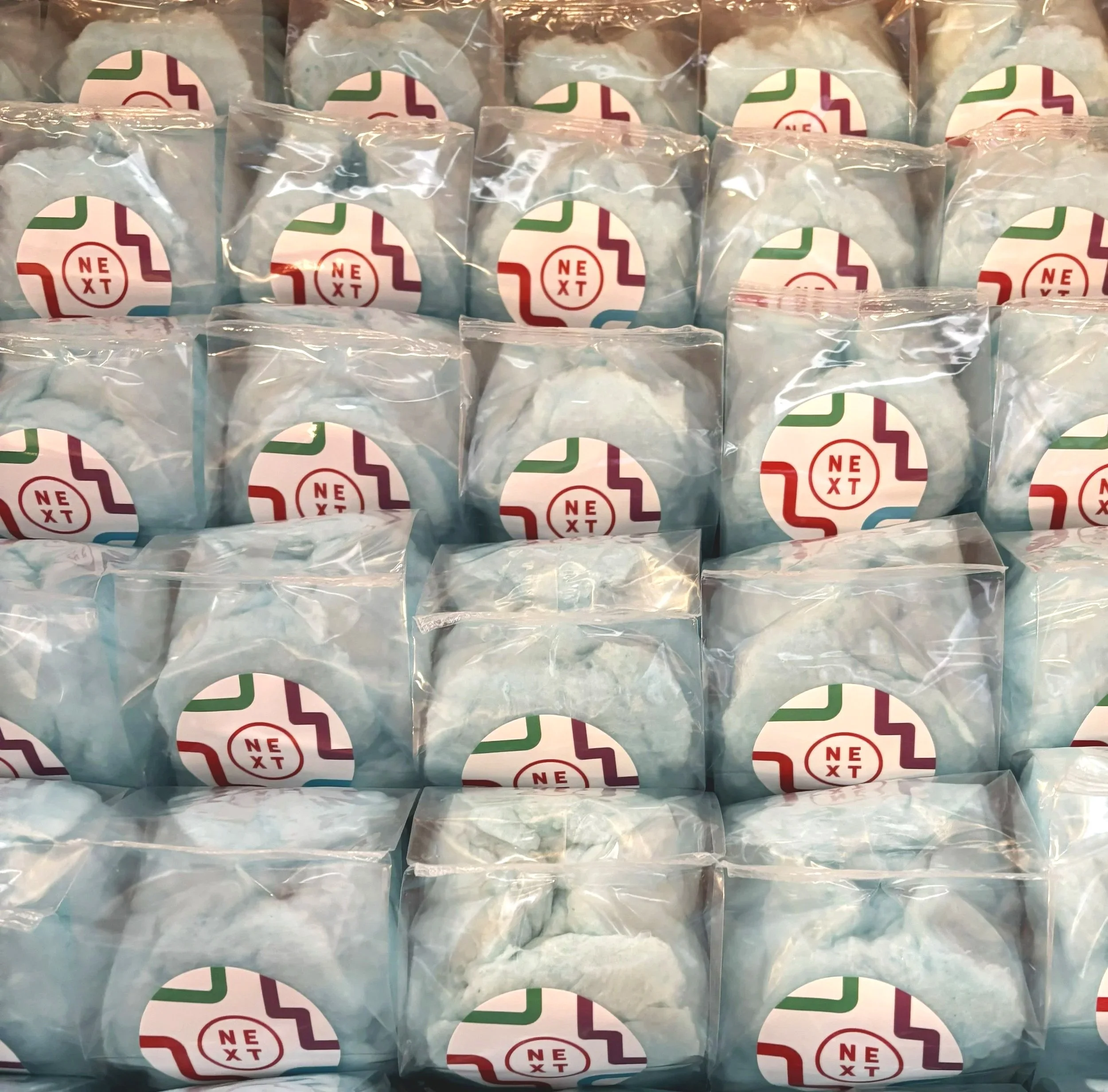 Multiple individually wrapped cotton candy bags with the Next logo on each wrapper, stacked on a shelf.