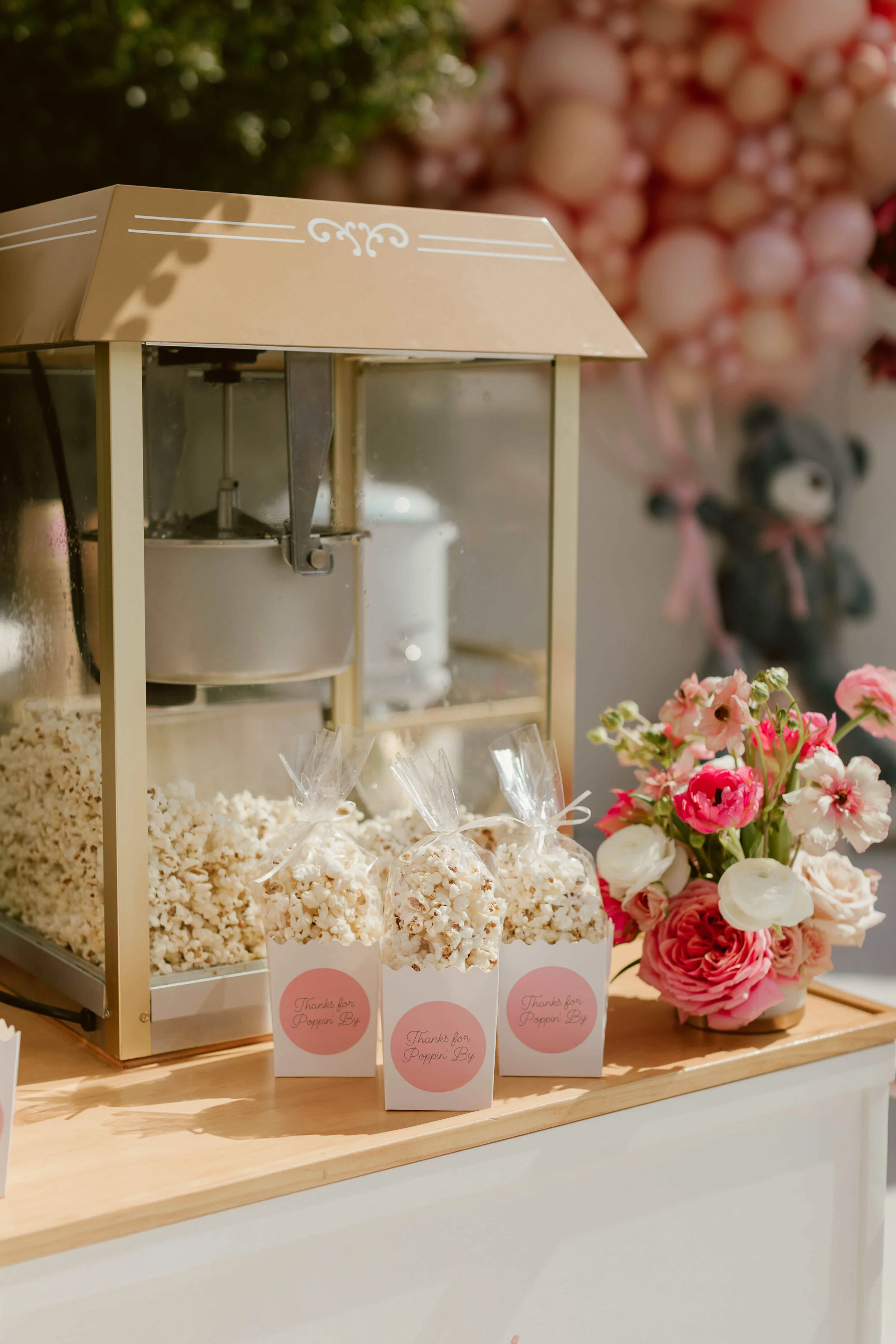3 boxed popcorn favors on a wooden table in front of a popcorn machine and next to a floral arrangement