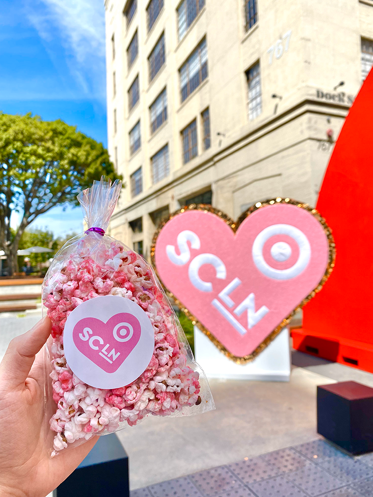 A hand holding a bag of pink and white popcorn with a sticker labeled 'SCLN' in front of a large building and a heart-shaped sign with the same label.