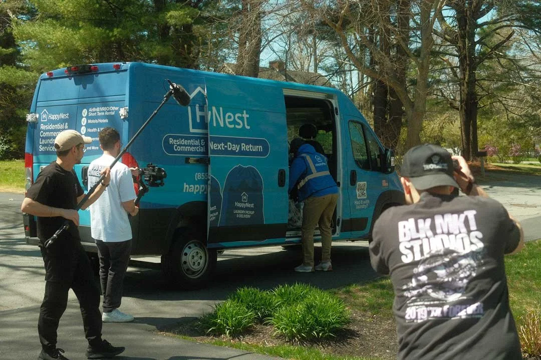 Filming crew filming an advertisement or documentary outside a blue HappyNest rental van with trees in the background.
