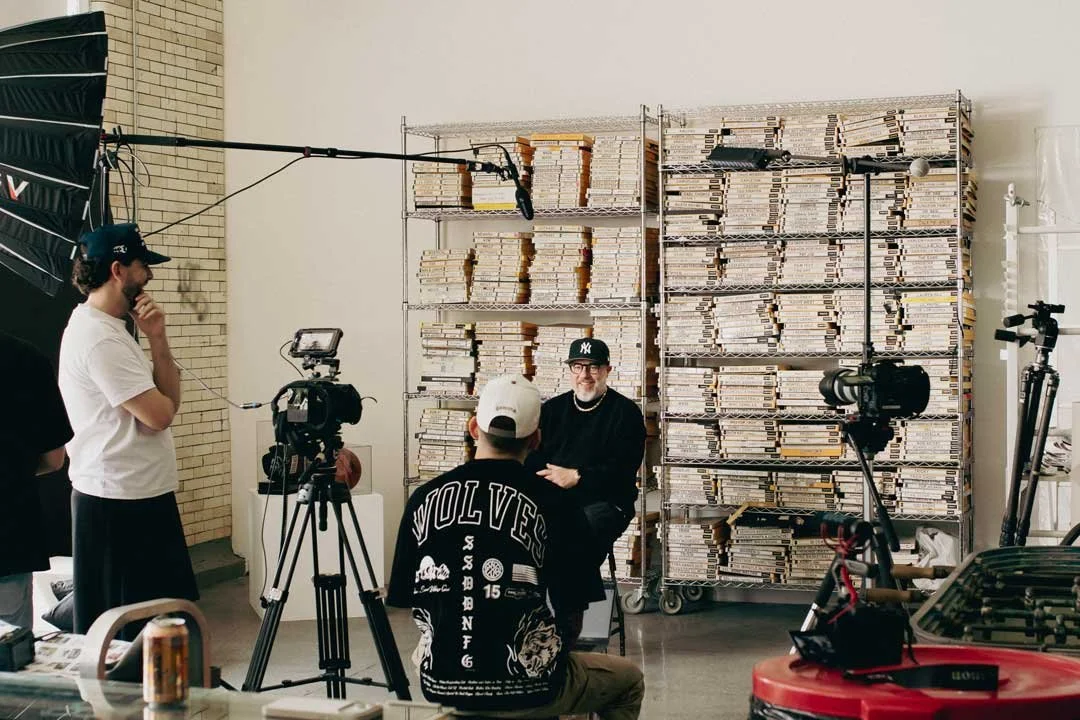 A man in black with a New York Yankees cap being interviewed by a film crew in front of large shelves filled with neatly stacked newspapers. The crew includes a camera operator, a person in a white t-shirt, and another with a cap that says "VAN". Equipment like cameras, a boom microphone, and tripods are visible.