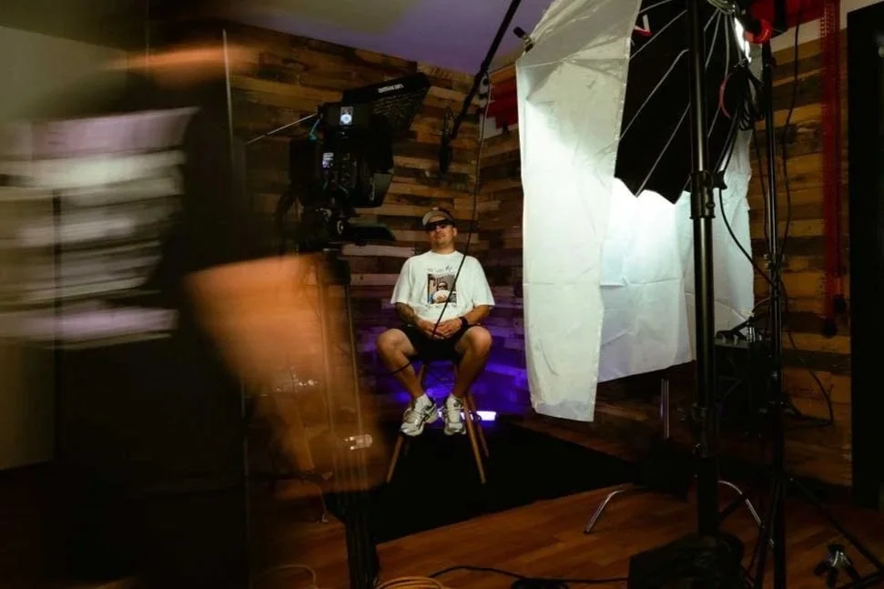 A person sitting on a stool in a photography or video studio with professional lighting and a backdrop, while a person moves past with a blurred effect.