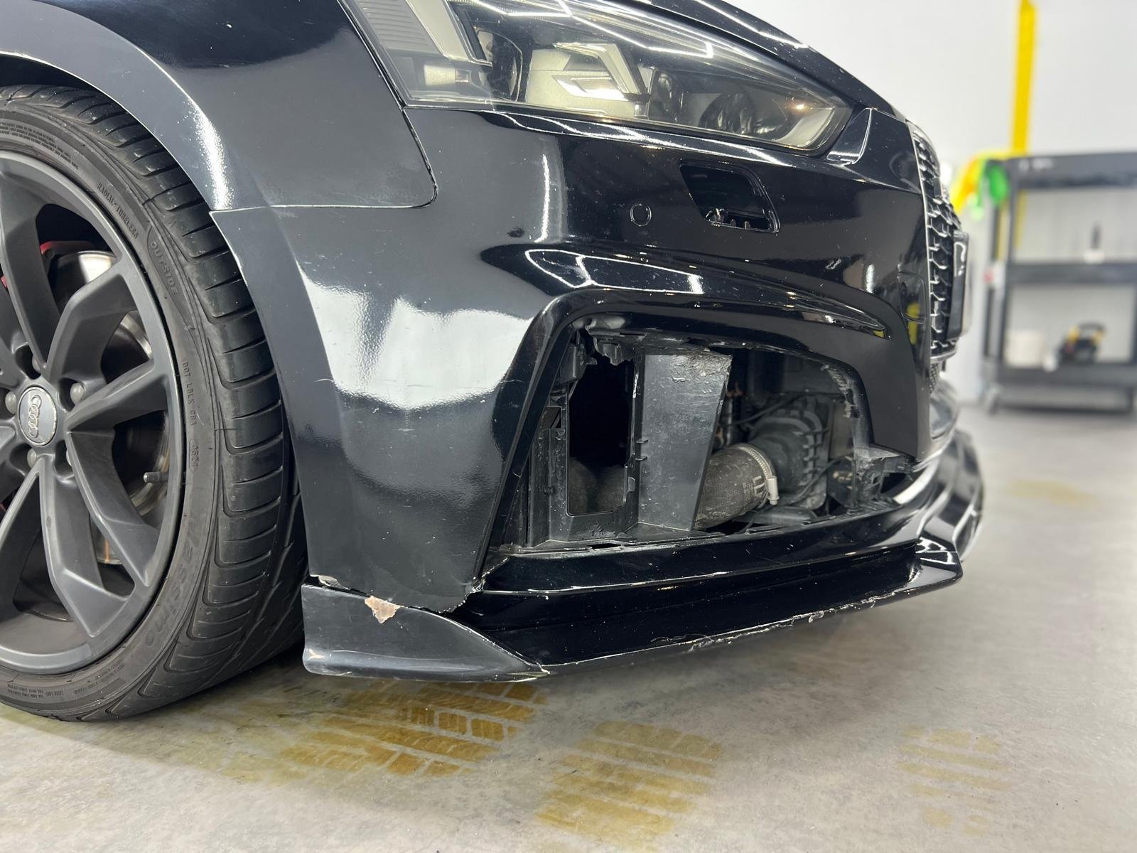 Close-up of a black car with damage to the front bumper, exposing internal components, in a garage or workshop.